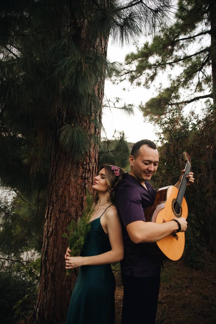 A couple poses romantically in a pine forest as they lean against a tree trunk, with the man holding an acoustic guitar.