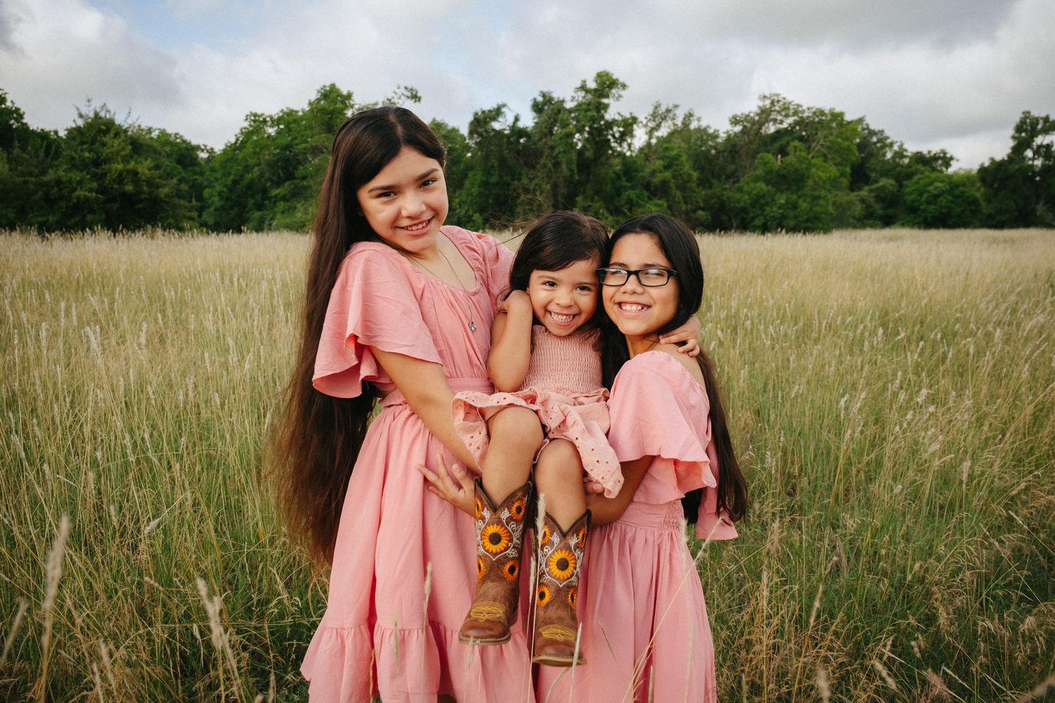Three girls in matching pink dresses pose in a golden field, two older sisters holding their younger sibling.