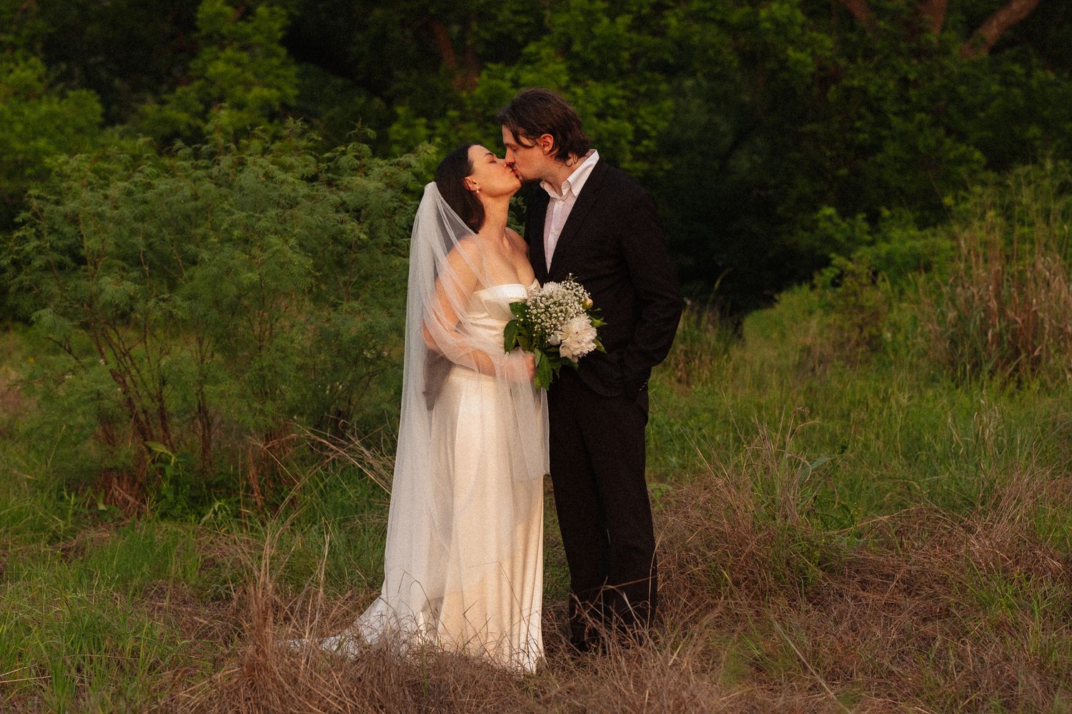 Bride and groom share a romantic kiss outdoors, bride wearing white gown with veil holding bouquet.