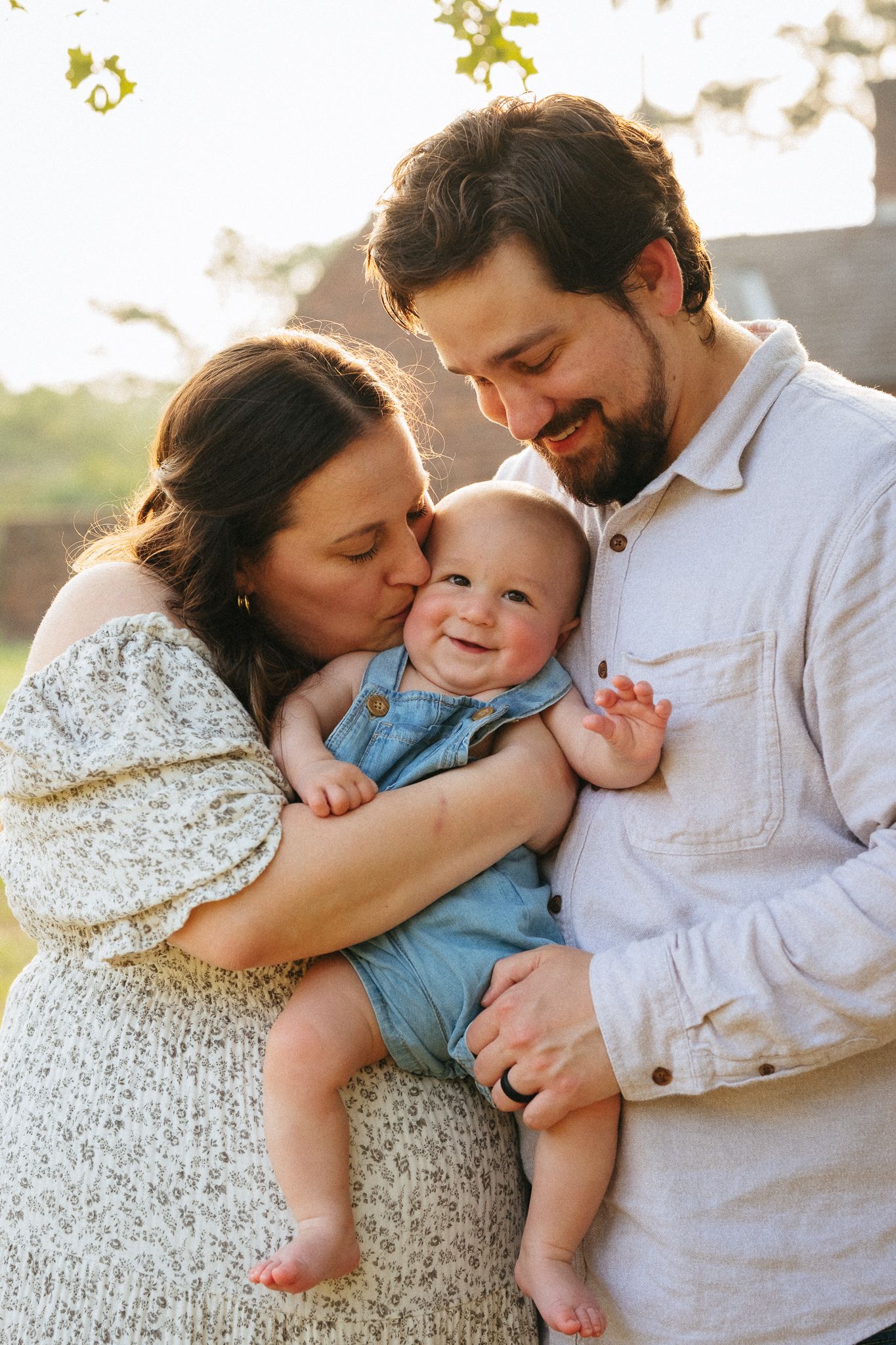 A happy couple holds their smiling baby outdoors in warm golden light, with the mother kissing the infant's cheek.