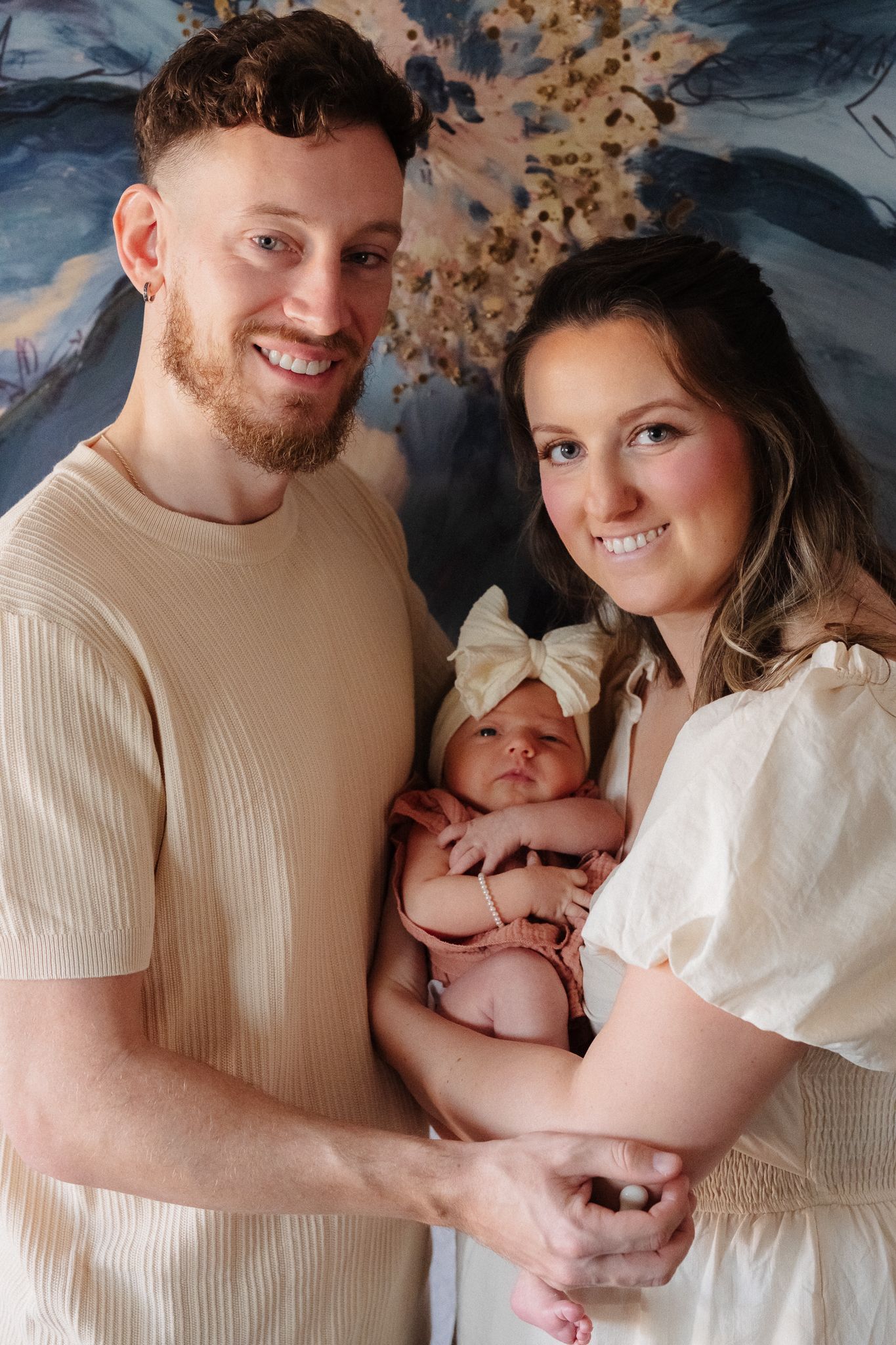 Smiling couple holding their newborn baby dressed in pink, posing together in front of an abstract painted backdrop.