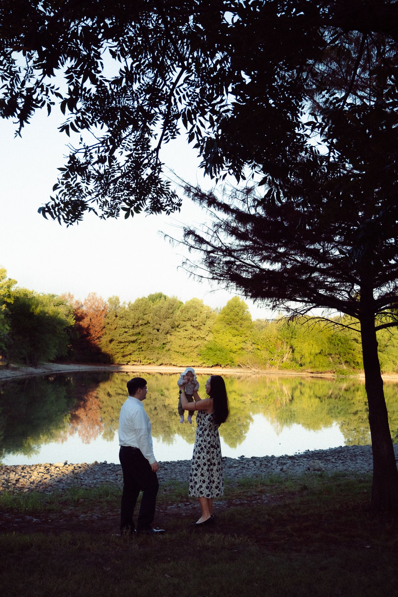 A couple stands in silhouette by a serene lake at sunset, framed by overhanging tree branches.