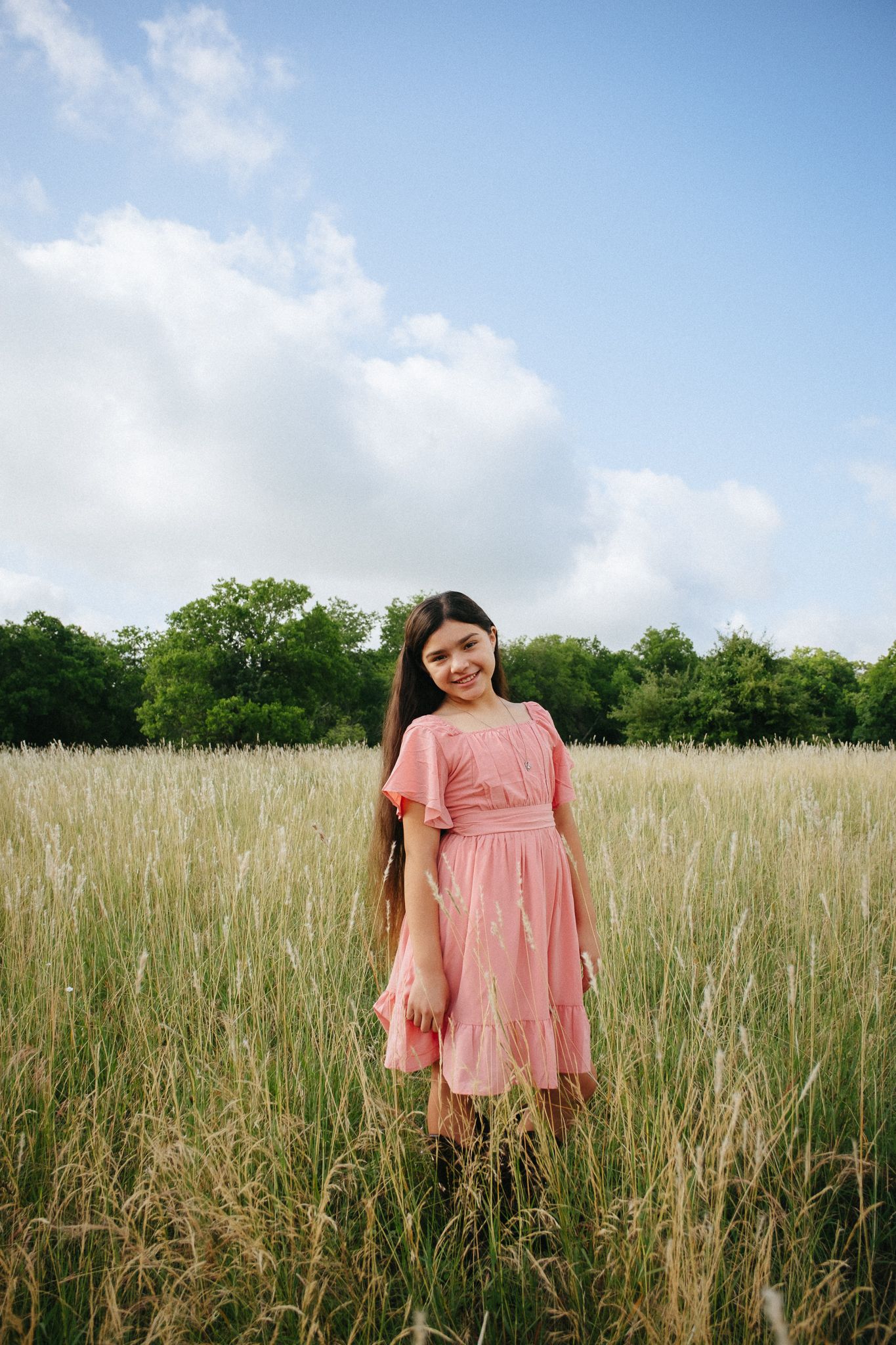 A person wearing a pink dress stands in a tall grassy meadow with trees and blue sky in the background.