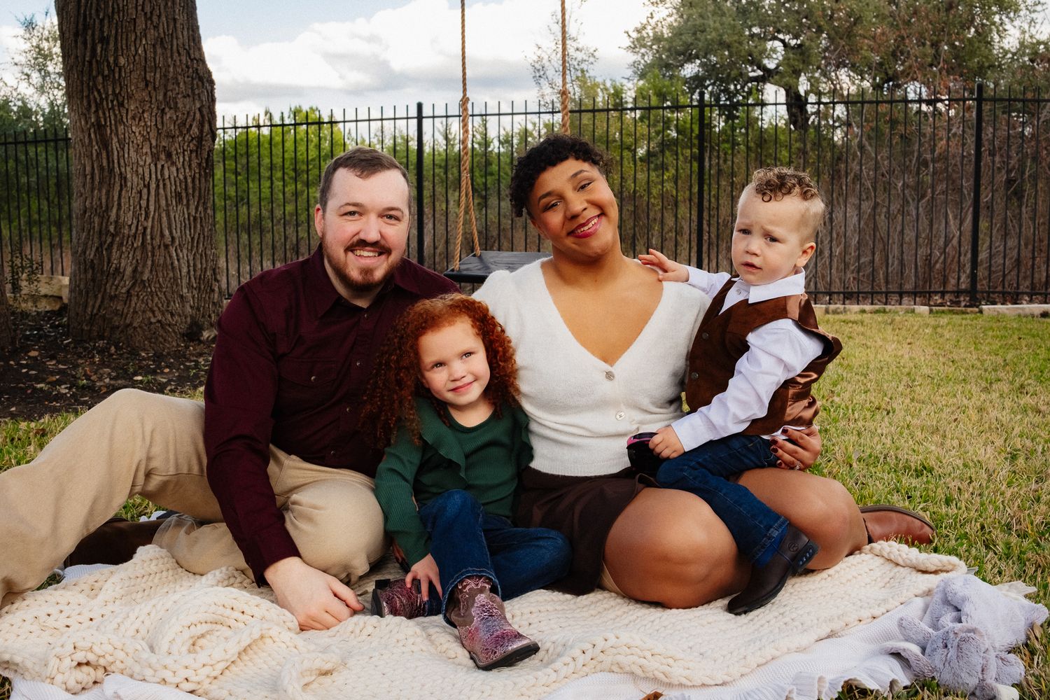 A family sits together on a blanket outdoors for a casual photo session in a backyard setting.