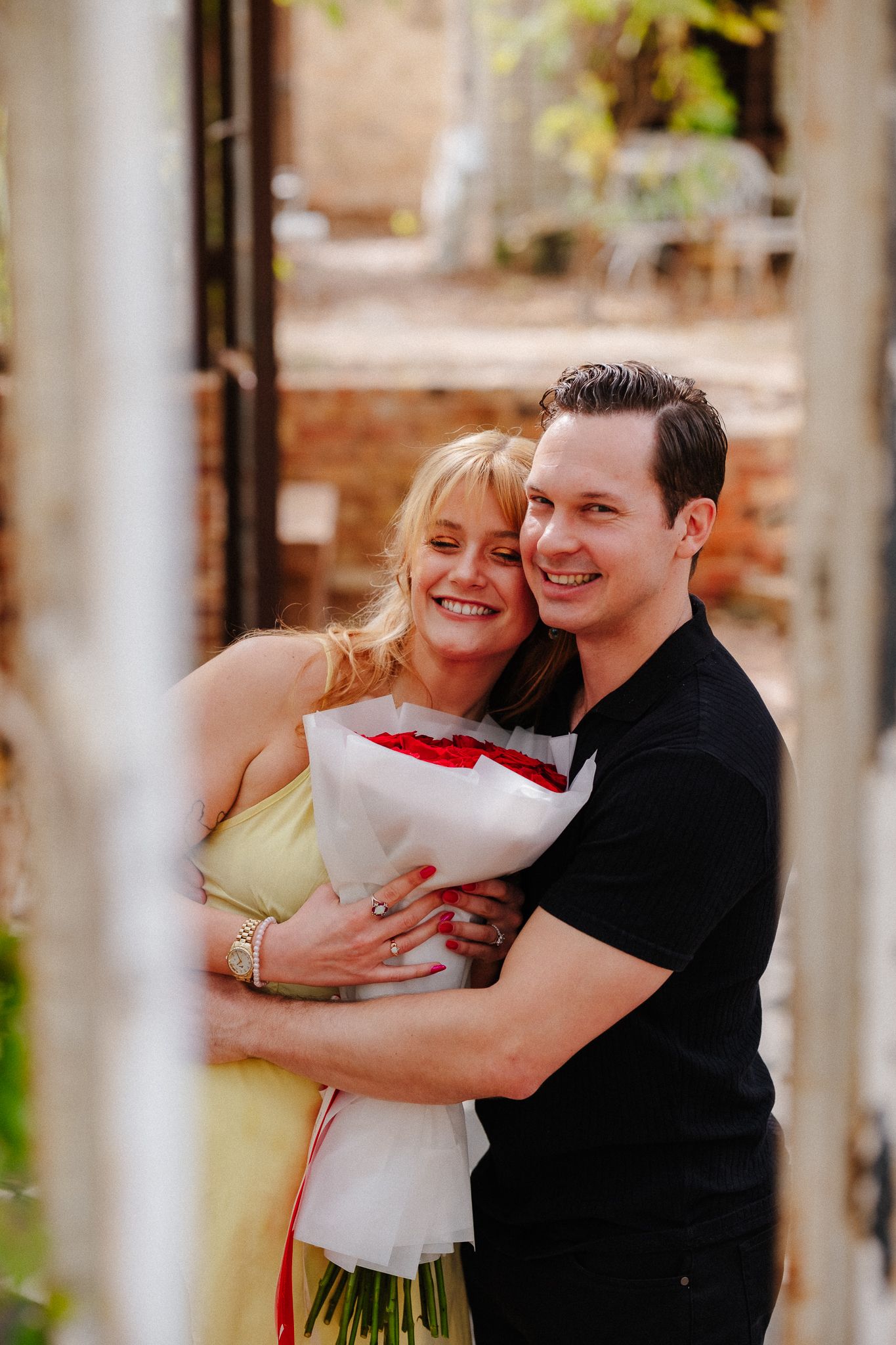 Smiling couple embracing outdoors, woman in yellow dress holding red roses bouquet, man in black t-shirt hugging her.