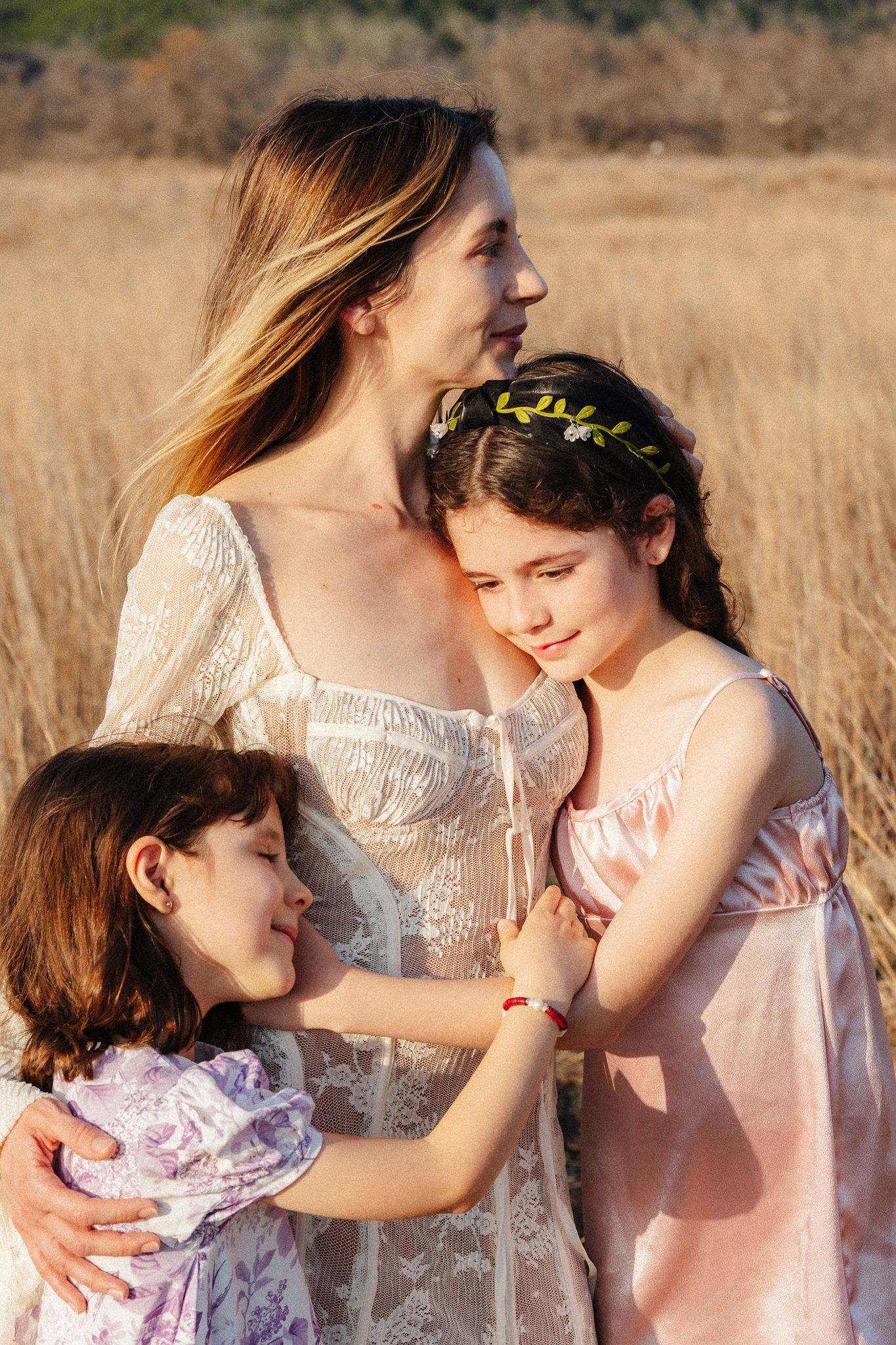 A mother and two daughters share a tender embrace in a golden field during warm sunset light.