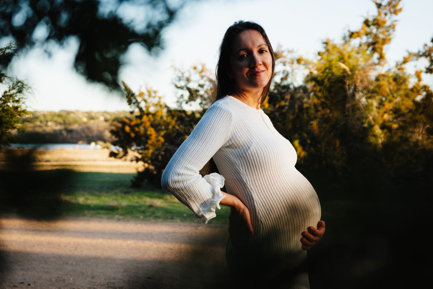Side view of expectant mother in white knit dress posing outdoors during golden hour.
