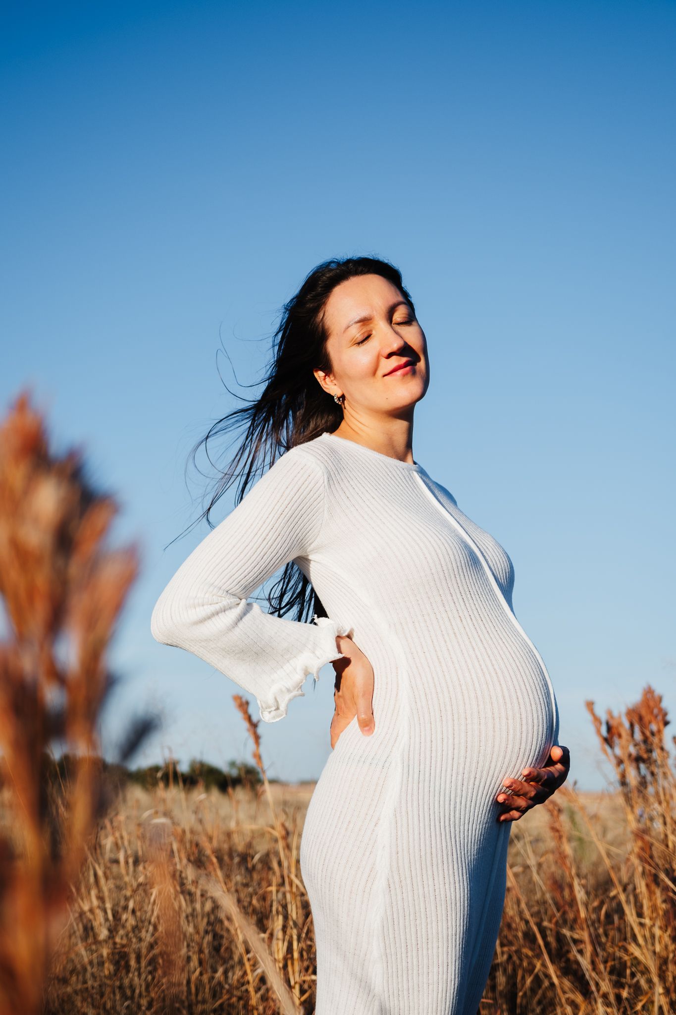 Pregnant woman in white fitted dress poses in field against bright blue sky during golden hour.