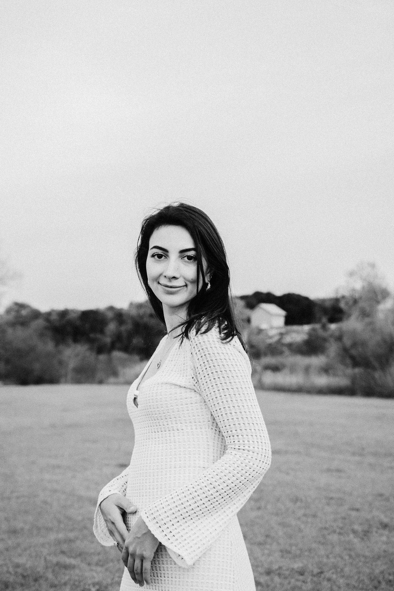 A woman in a white crocheted dress poses in a field with a soft smile in black and white photography.