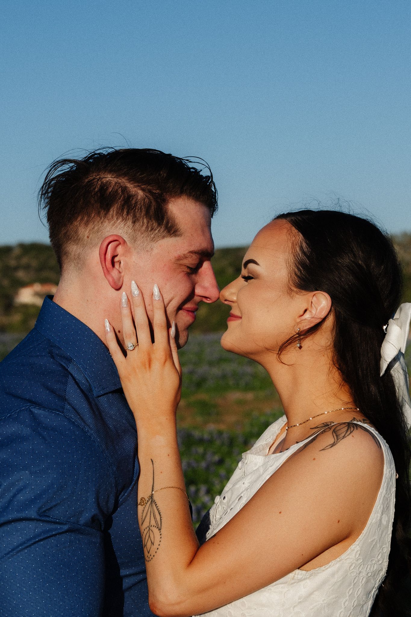 A bride and groom share a romantic almost-kiss moment outdoors, with bluebonnets and blue sky in the background.