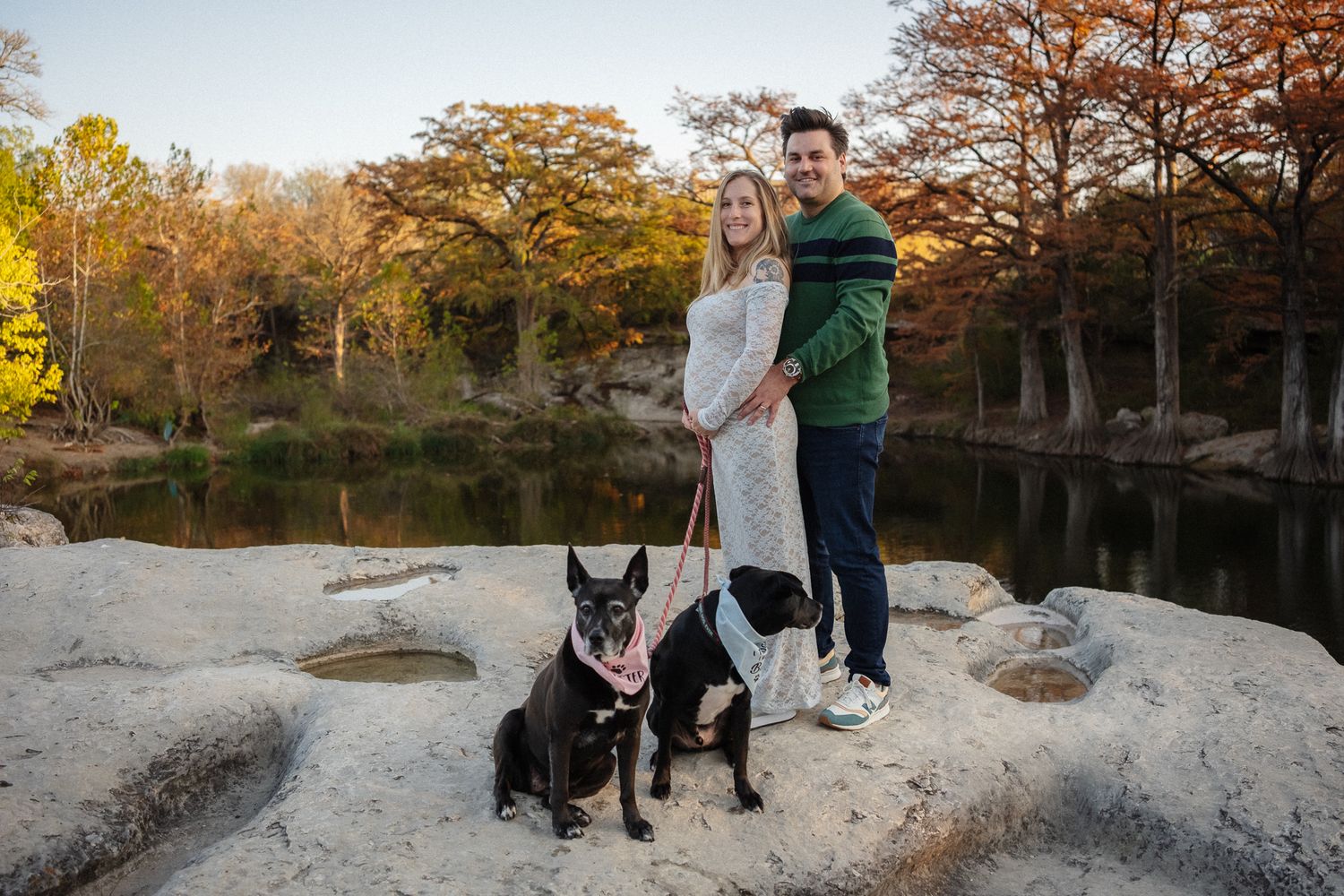 A person and a black dog stand together on a rocky outcrop during autumn at sunset.