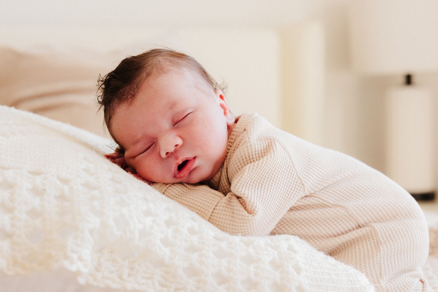 Peaceful sleeping newborn baby wearing white knit clothing rests on soft cream-colored bedding.