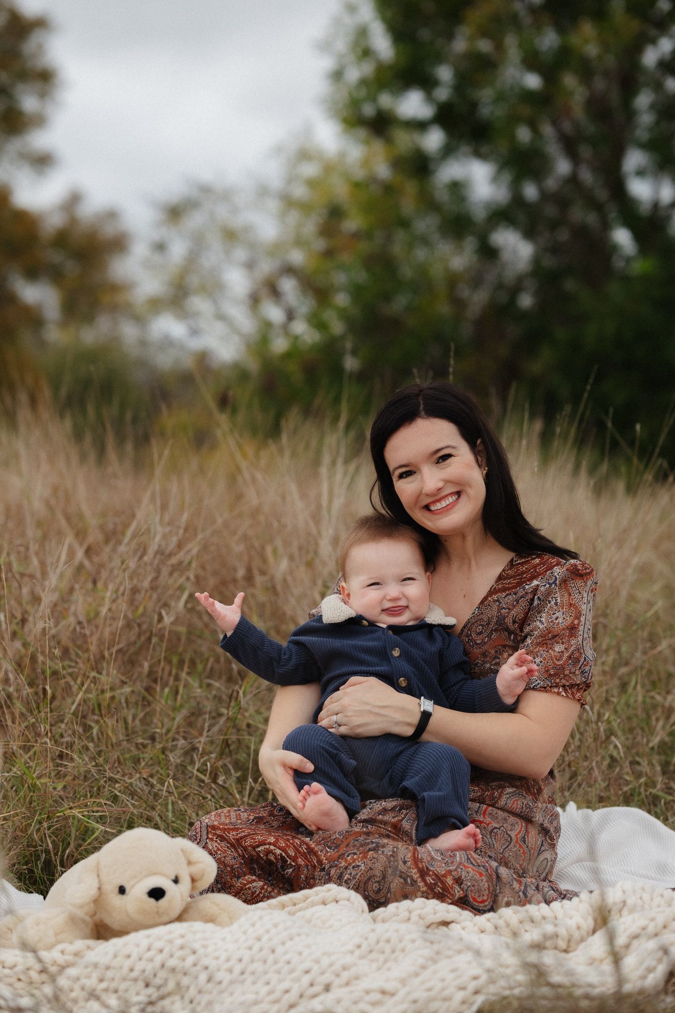 Mother and baby sitting on cozy knit blanket in tall grass during outdoor photo session.