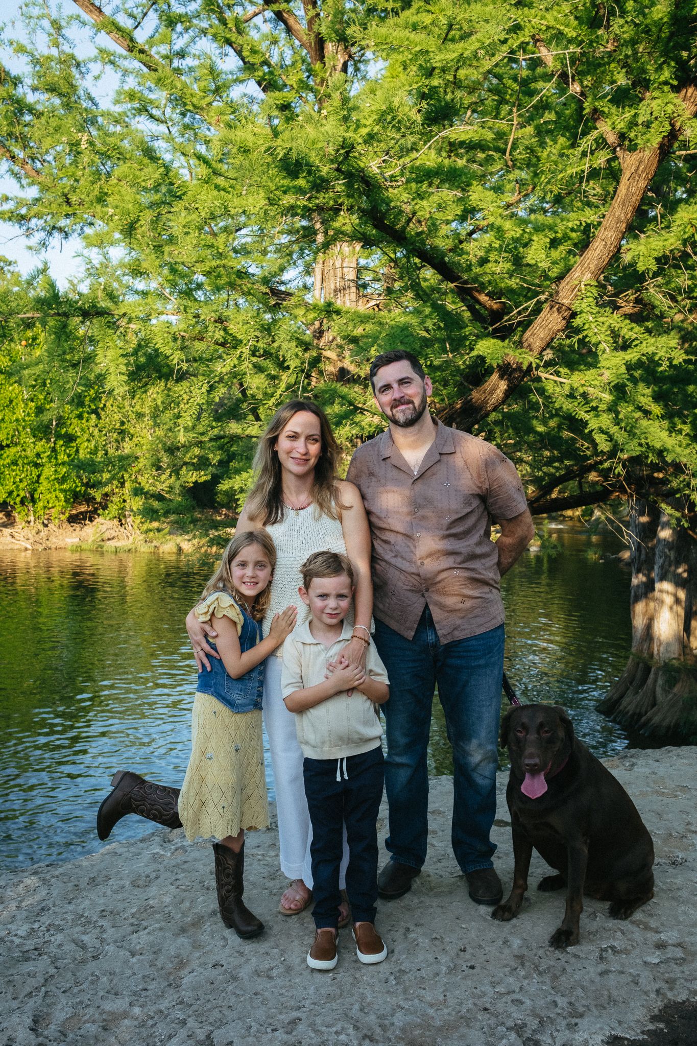 Family of four with a chocolate Labrador posing by a river surrounded by lush green trees.
