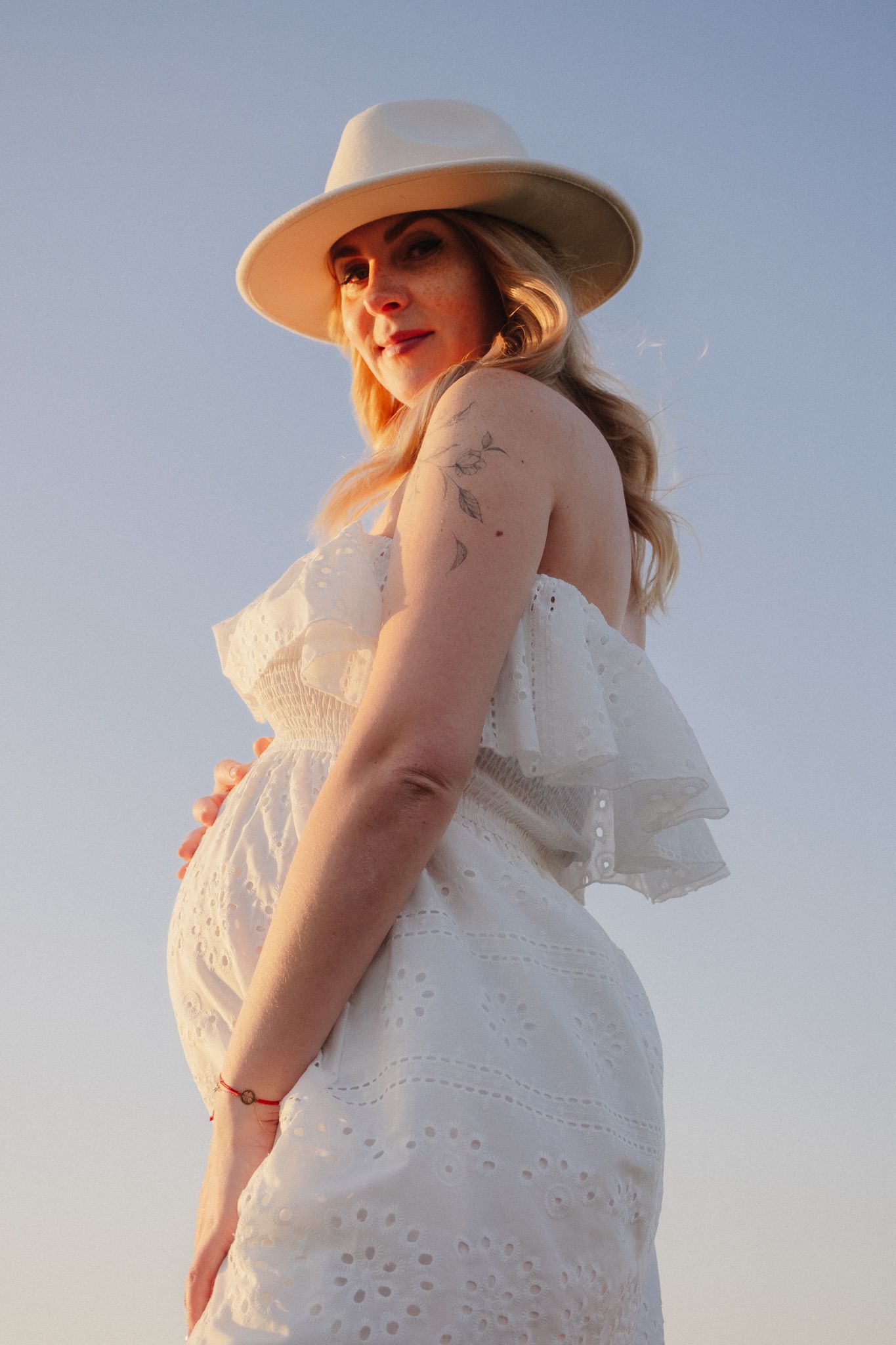 Pregnant woman in white eyelet dress and wide-brim hat cradling baby bump at golden hour.