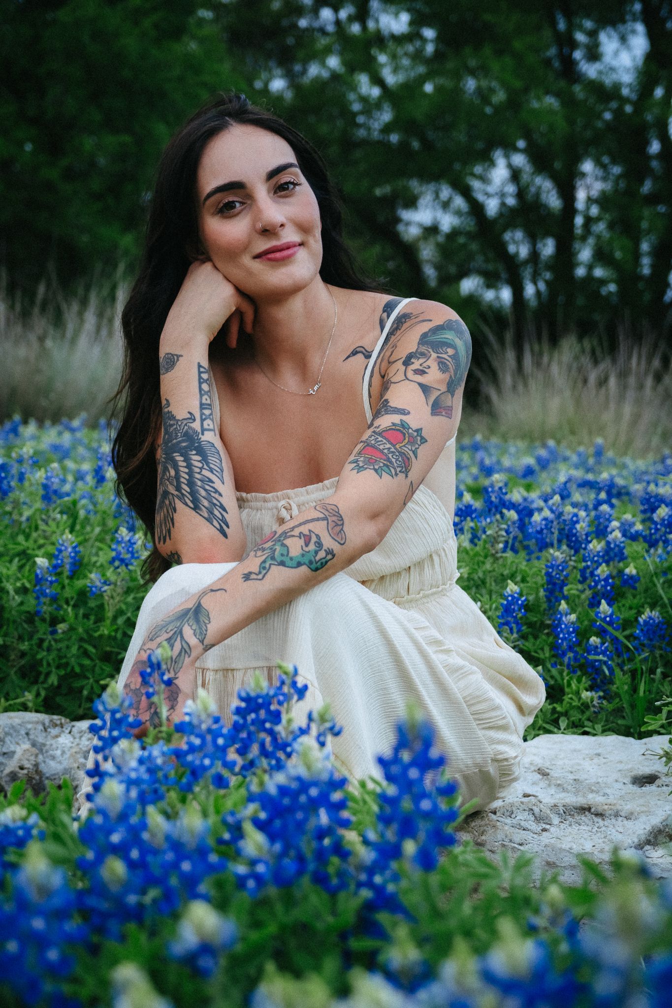Person in white dress poses among vibrant bluebonnet flowers with visible arm tattoos and dark long hair.