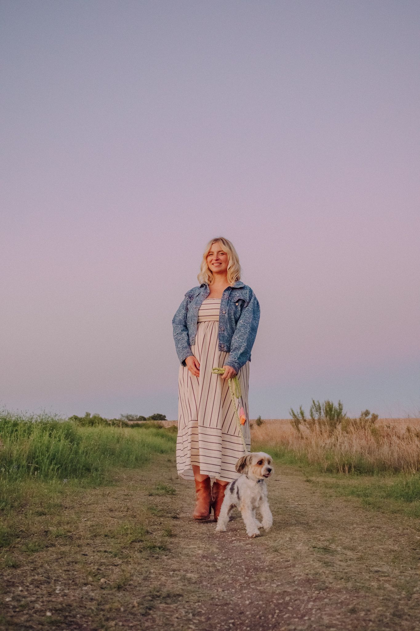 A person in a denim jacket and striped dress walking with a white dog at sunset in a grassy field.