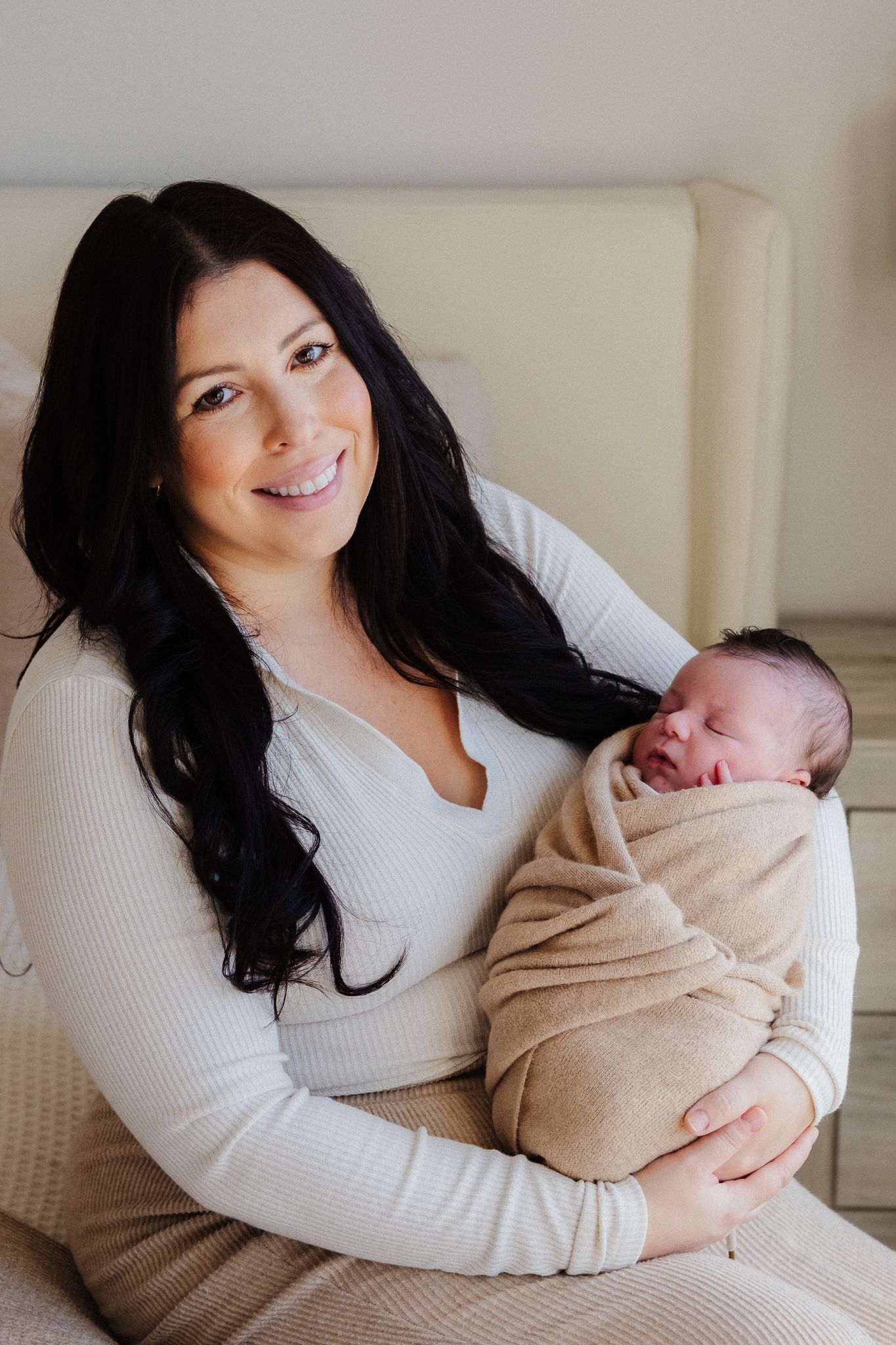Mother in white sweater holding and bonding with newborn baby wrapped in beige blanket in bright studio setting.
