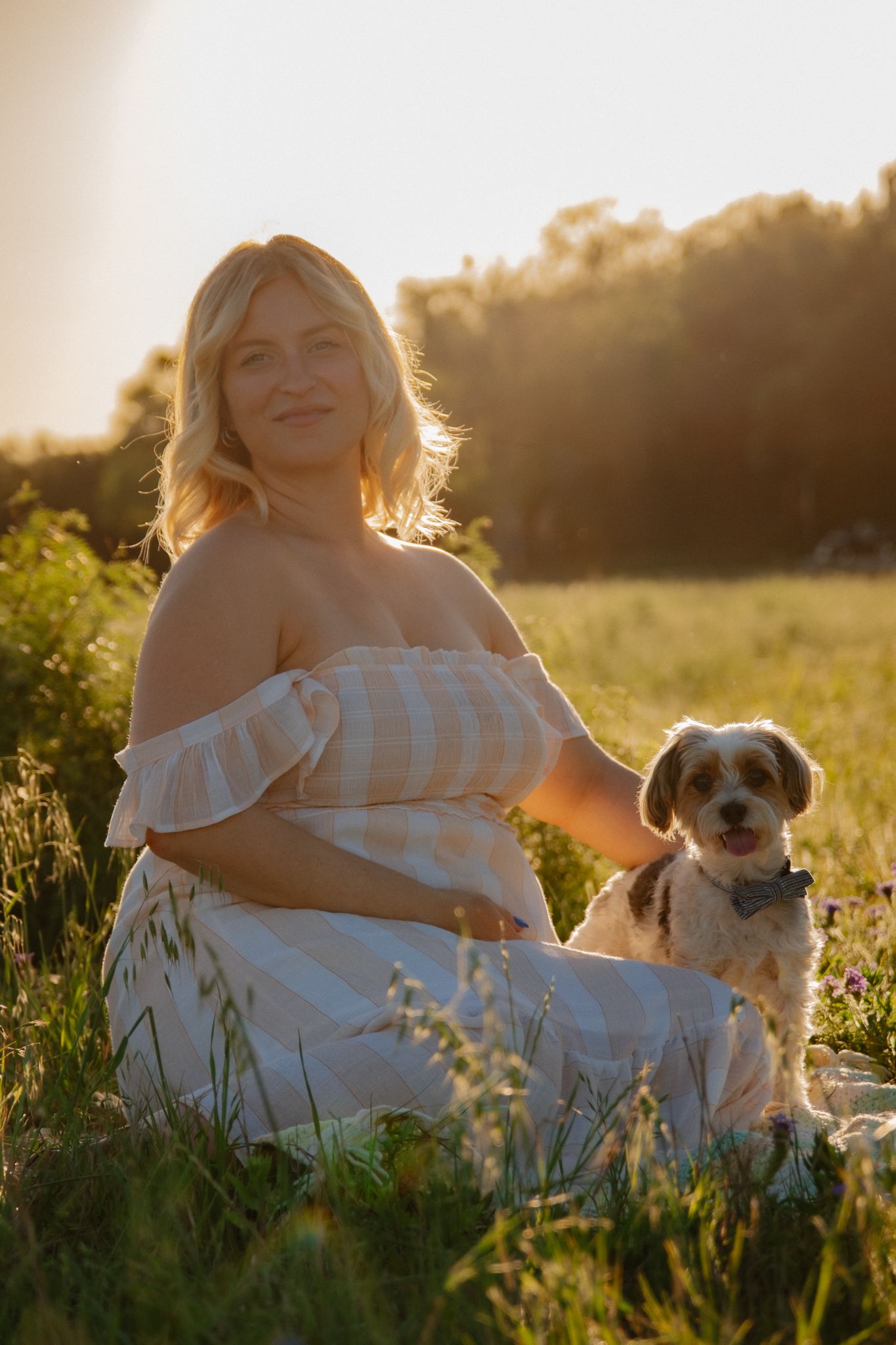 A person in a white dress sits in a sunlit meadow at golden hour with their small dog beside them.