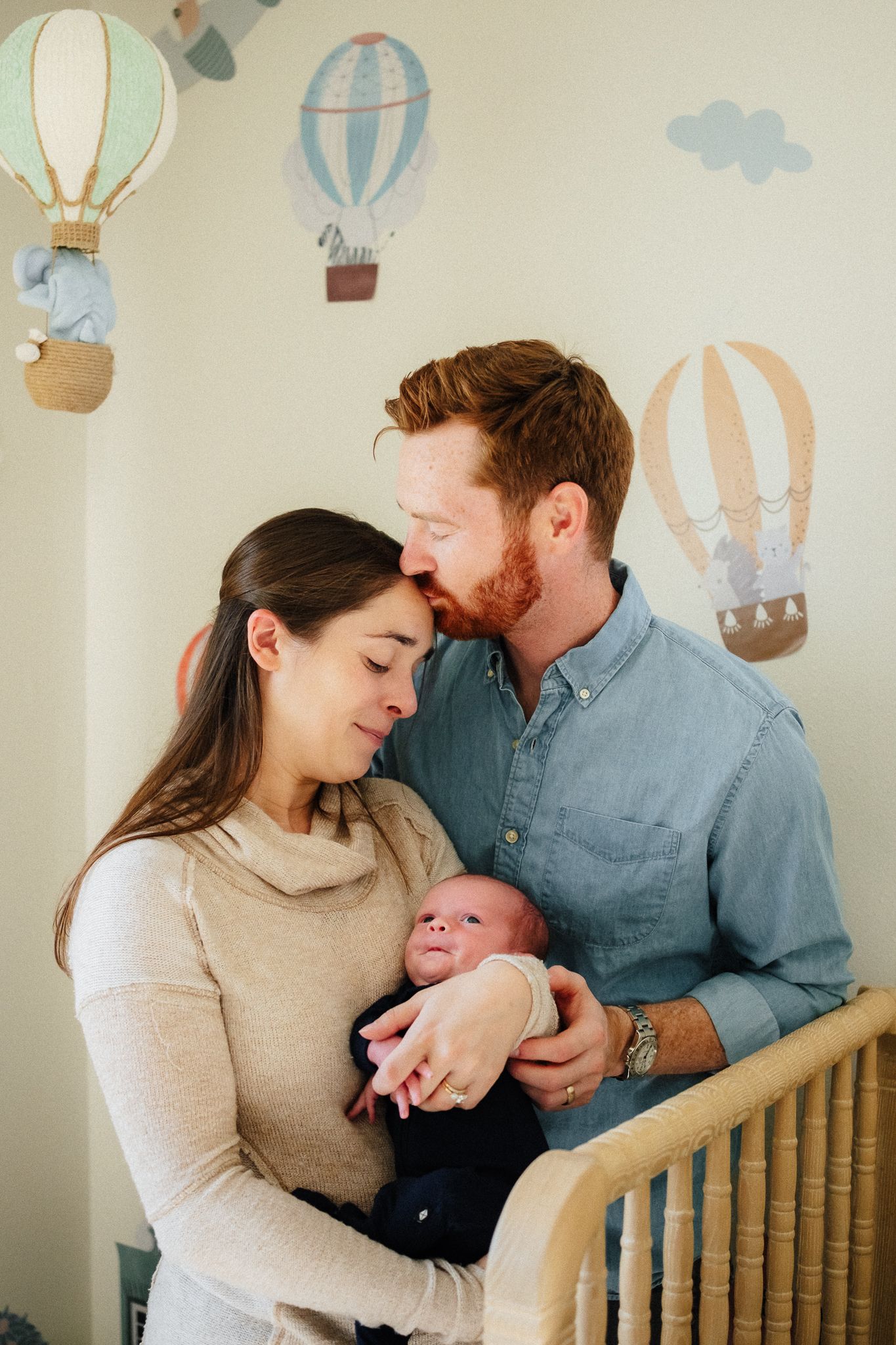 Parents share an intimate moment with their newborn in a nursery with whimsical wall decorations.