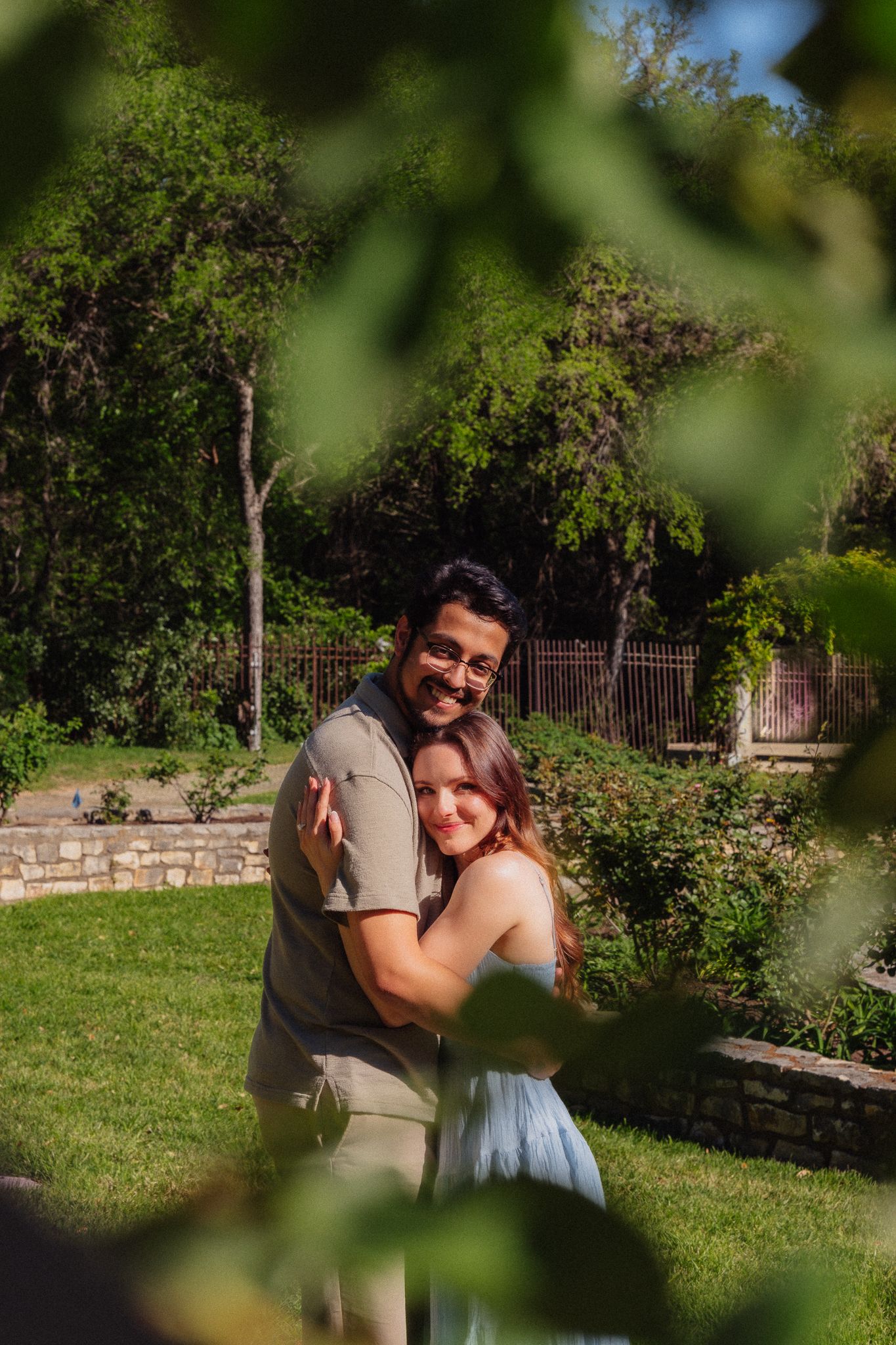 Couple embracing and smiling in a lush green garden with trees and stone walls in the background.