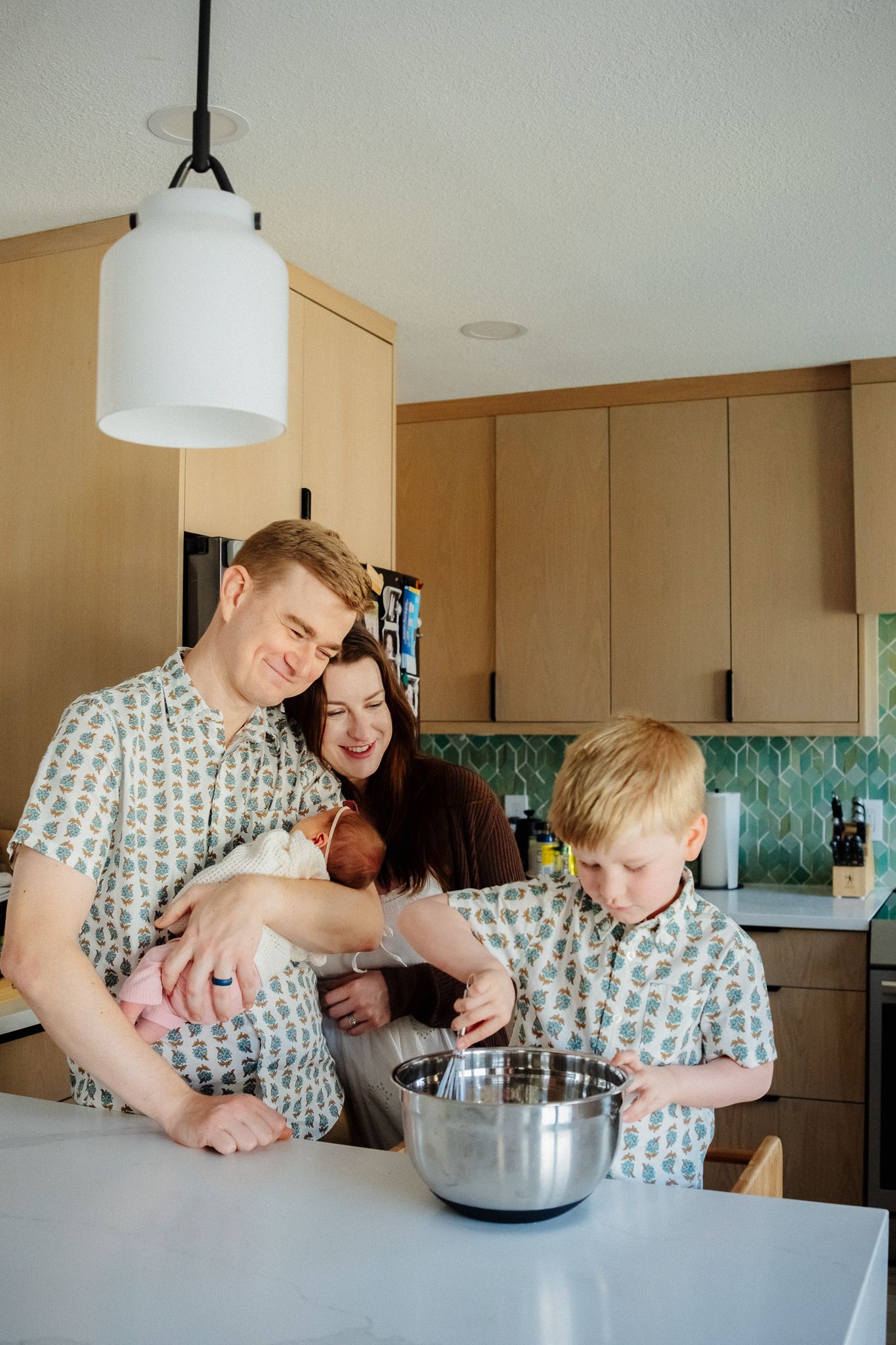 Family of four baking together in a modern kitchen with wood cabinets and teal backsplash, parents and children around a mixing bowl.