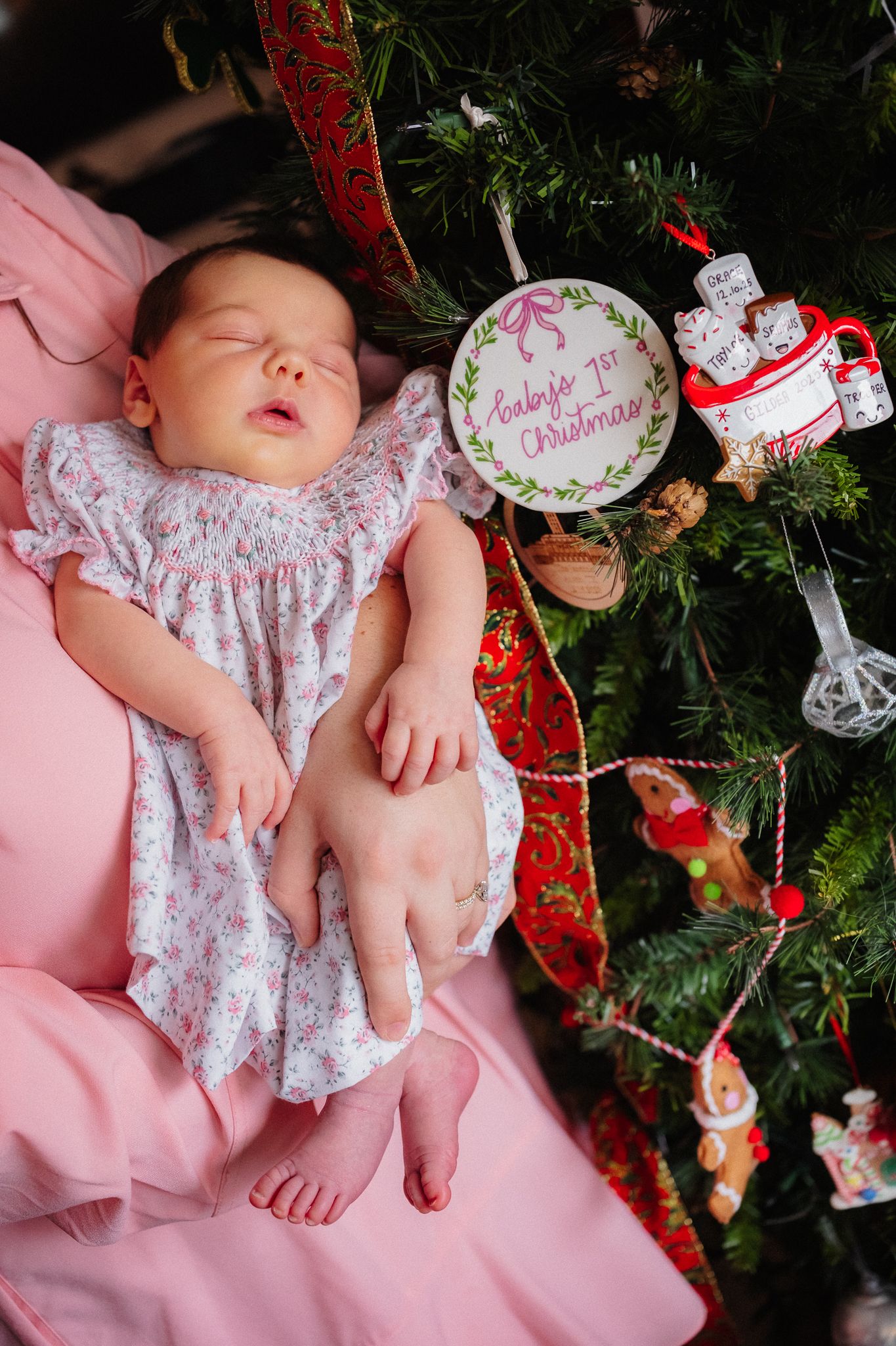 Sleeping newborn baby in floral dress beside Christmas tree with Baby's 1st Christmas ornament on pink blanket.