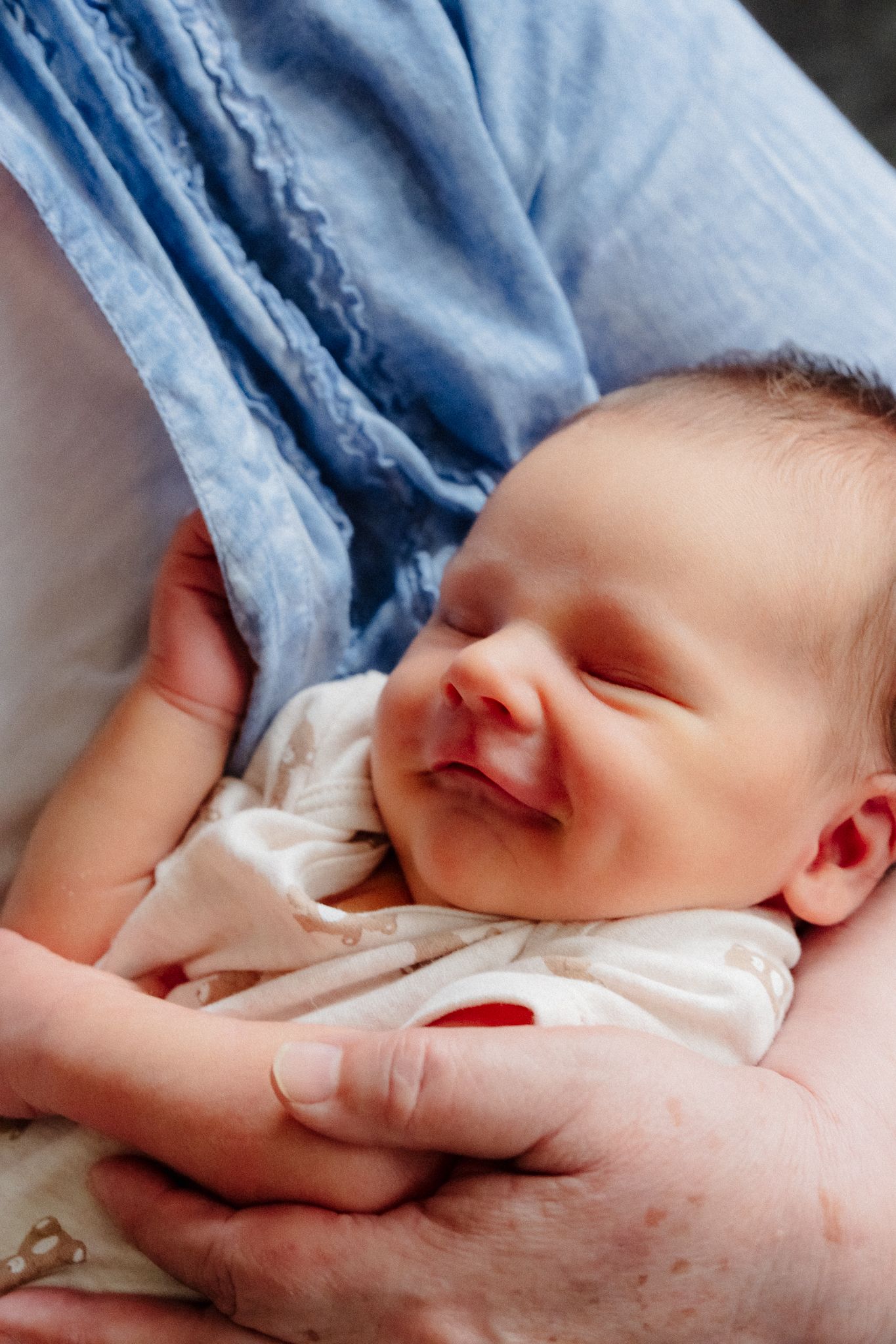 Newborn baby sleeping peacefully in caregiver's arms, wearing white onesie, with a gentle smile on face.