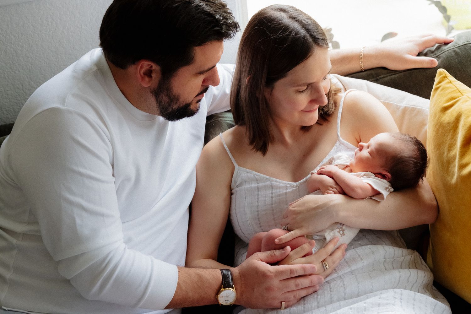 New parents sitting on a couch together, lovingly gazing at their newborn baby held in the mother's arms.