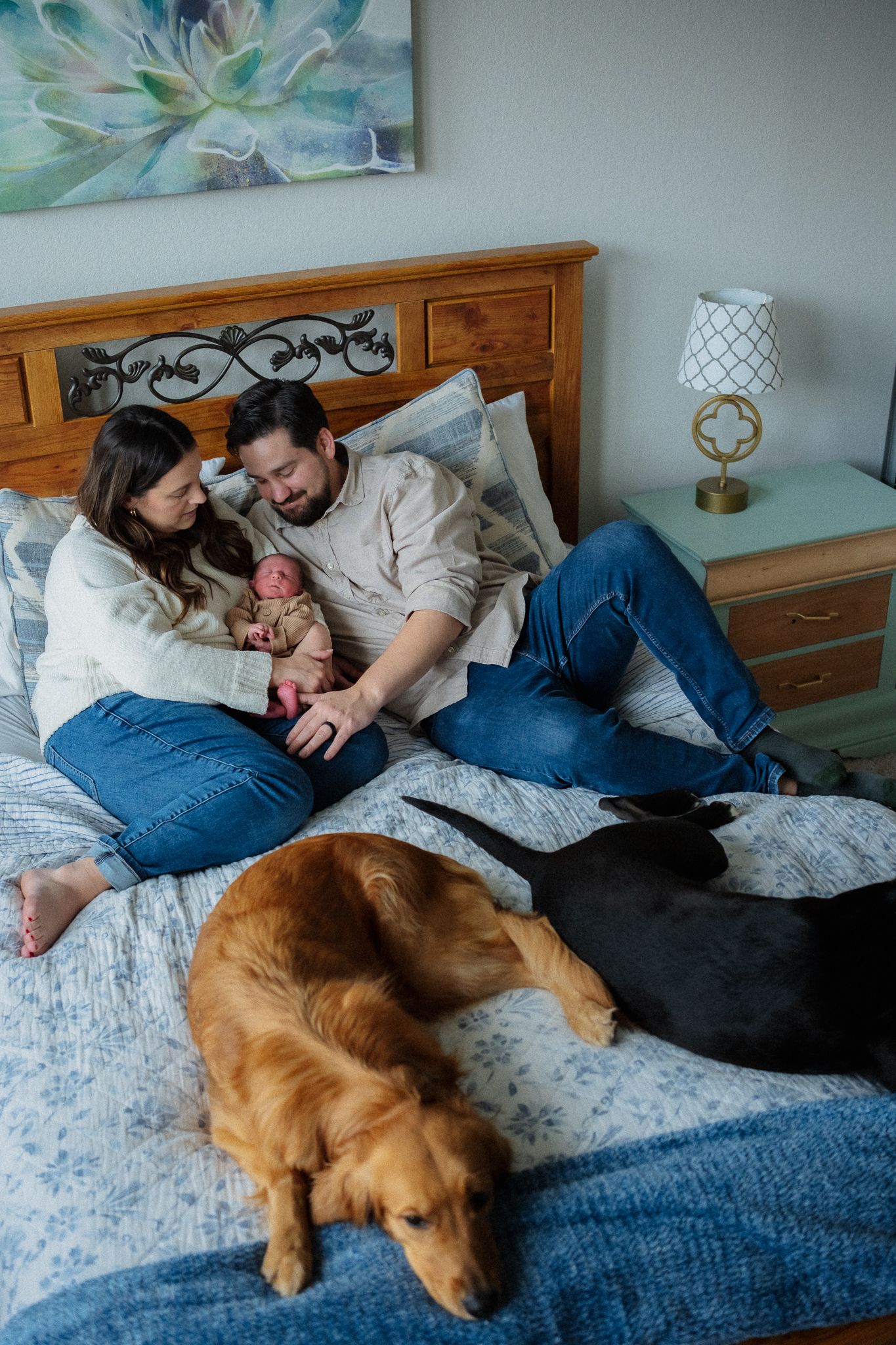 A couple relaxes on a blue bedspread with their golden retriever dog laying at the foot of the bed.