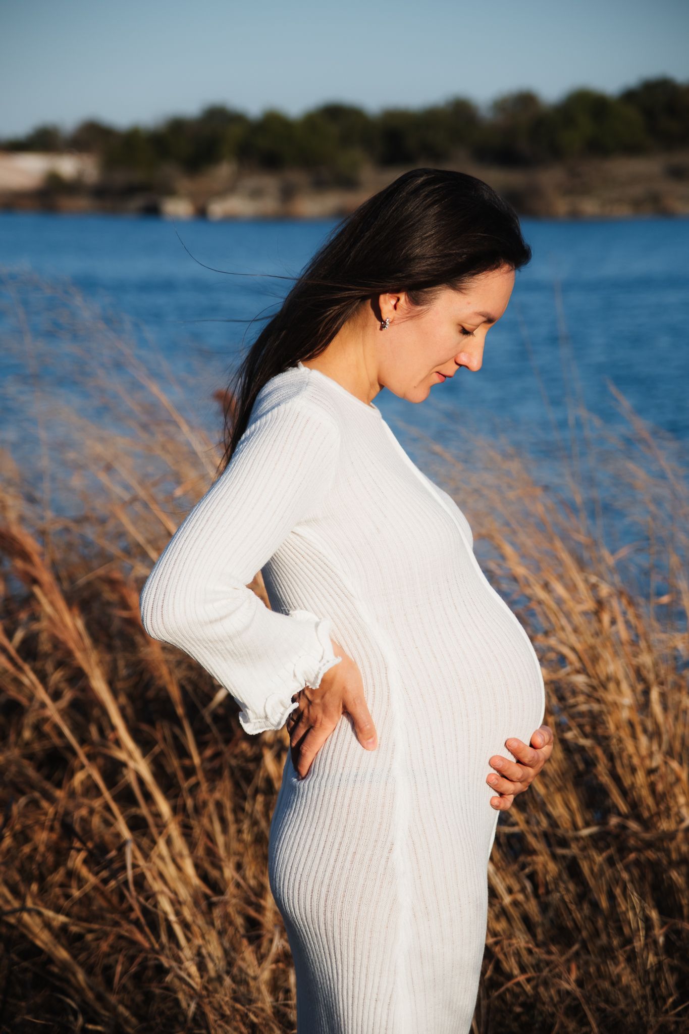 Expectant figure in white ribbed dress stands beside water at golden hour among dry grasses.