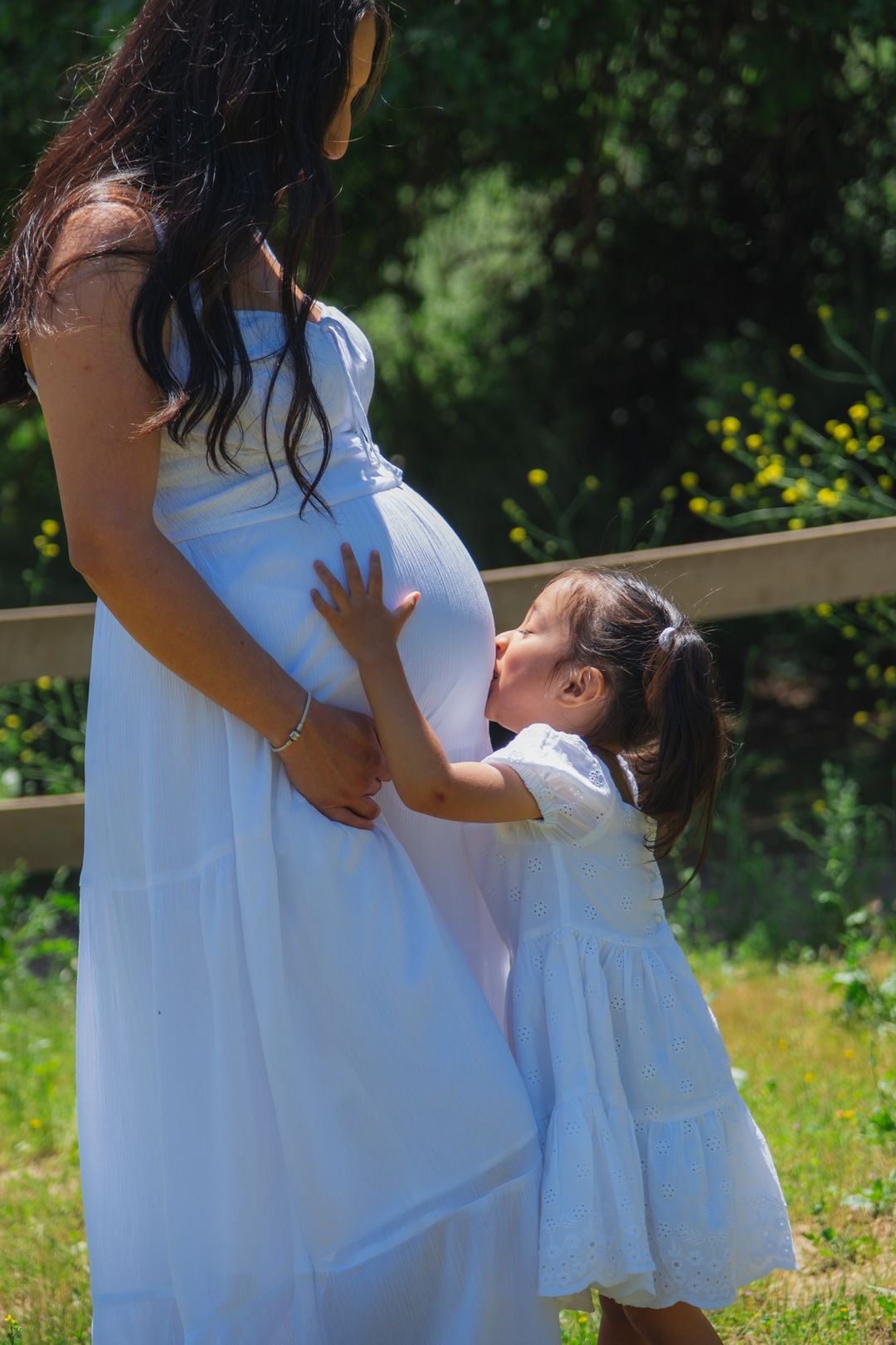 Tender moment in a garden setting as a child kisses a pregnant belly while both wear matching white dresses.