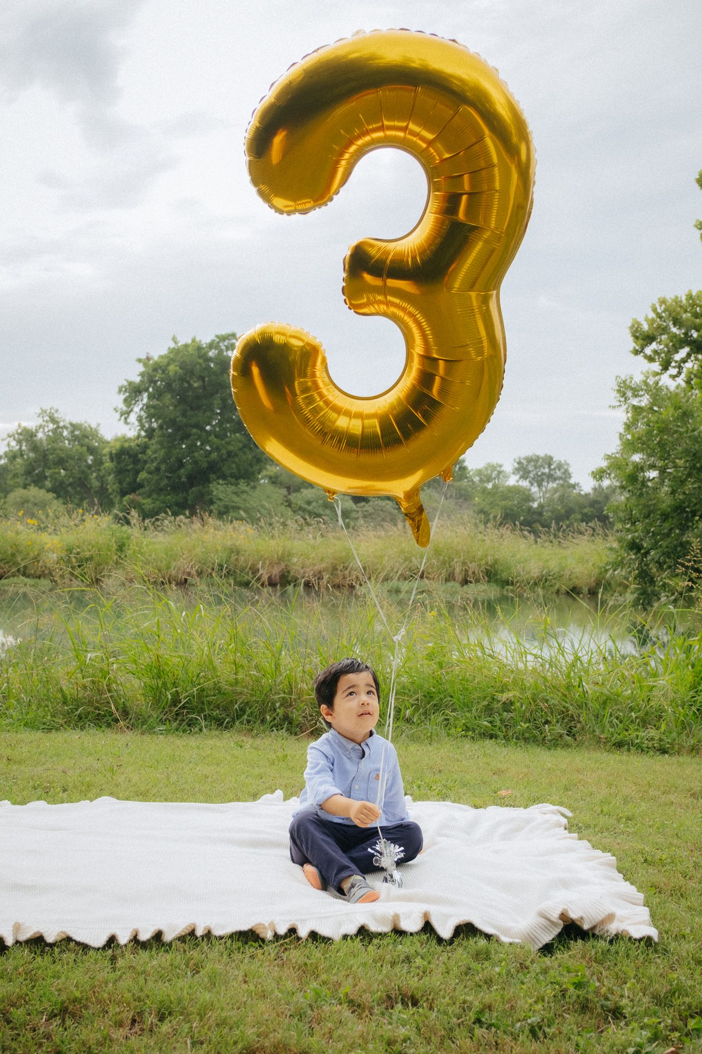A photo series on white blanket outdoors with gold number 3 balloon floating in a grassy field on sunny day.