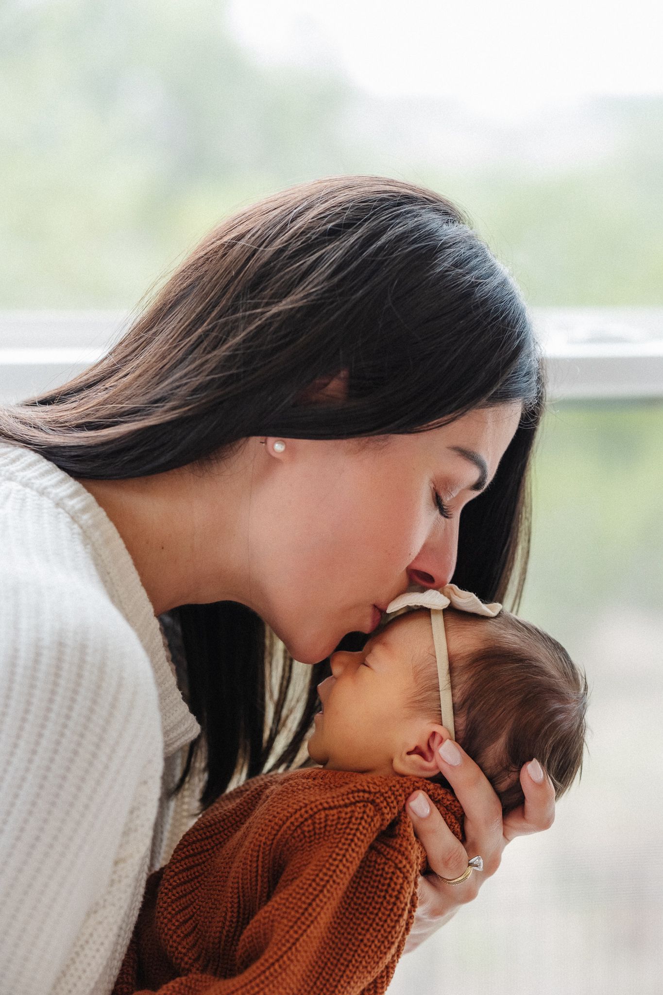 Mother tenderly kissing her newborn baby wrapped in rust orange knit blanket near a bright window.