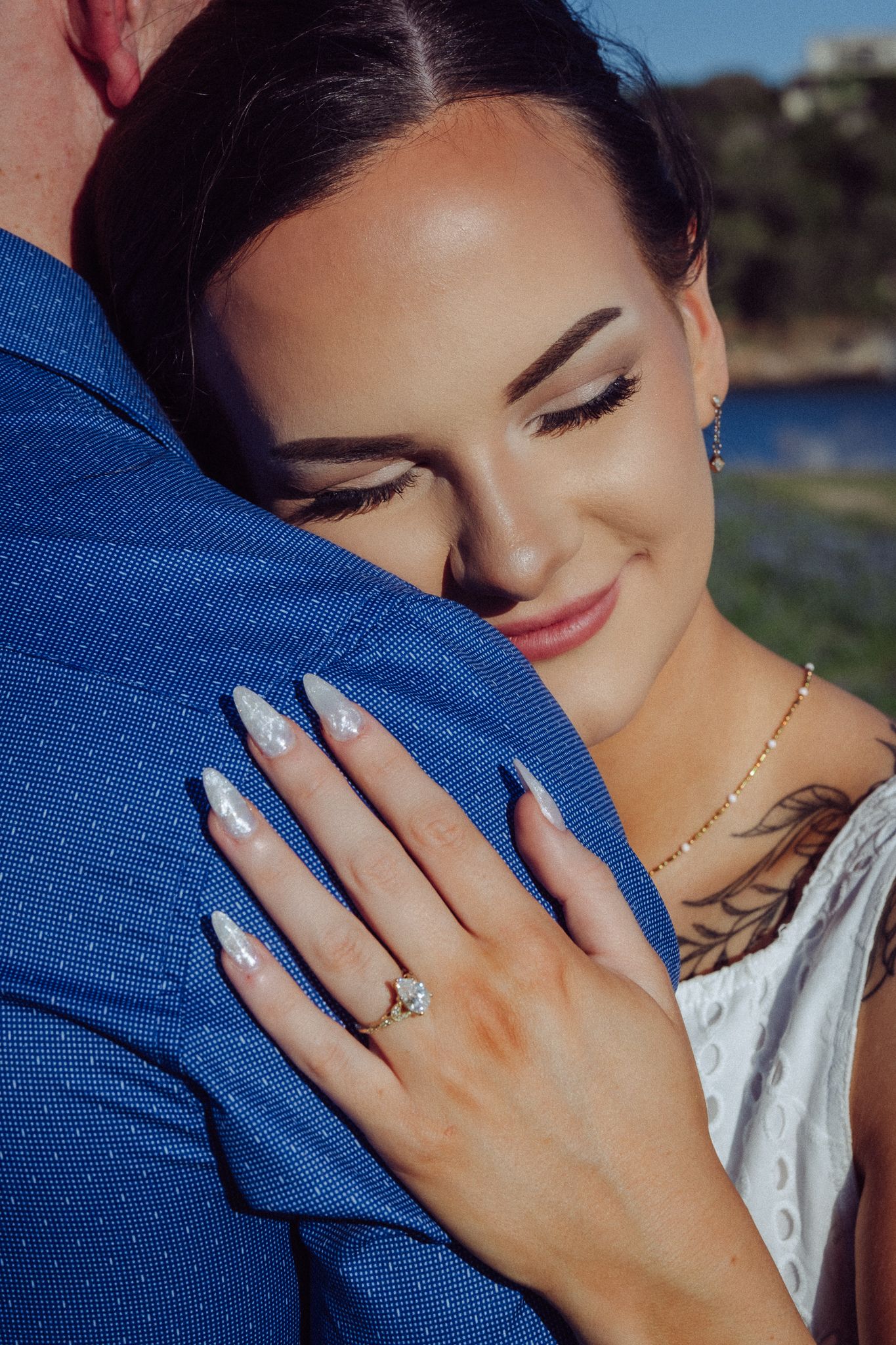 Smiling bride with long nails and engagement ring embracing groom in blue suit outdoors near water.