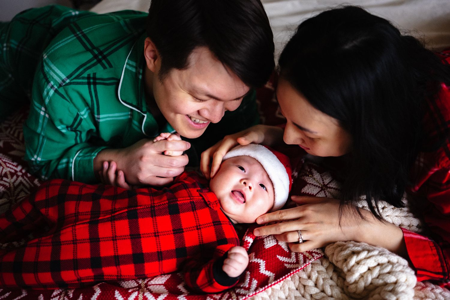 Two parents in Christmas pajamas lovingly gaze at their newborn baby wearing a Santa hat on a red plaid blanket.