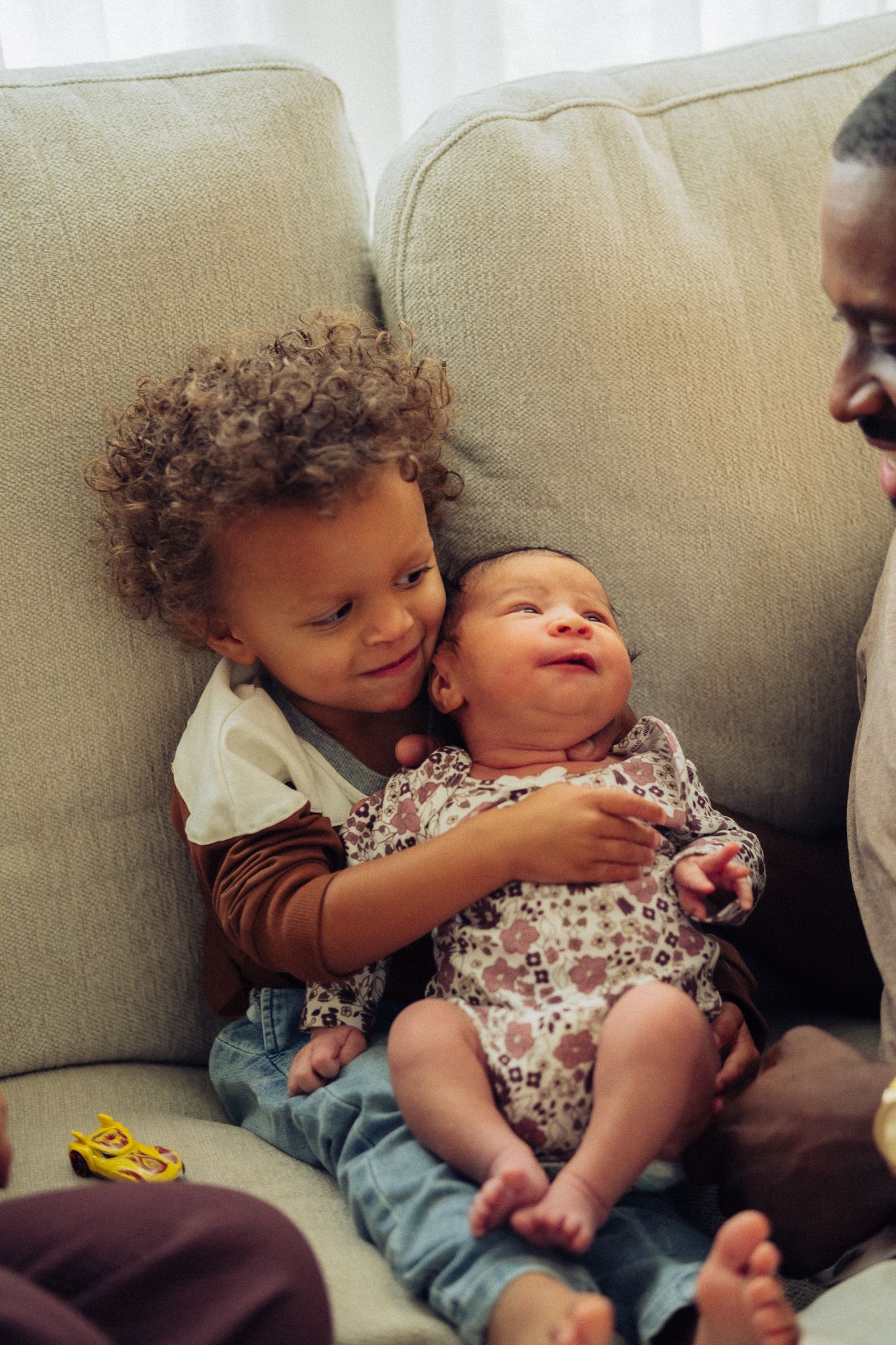 A toddler with curly hair gently holds a smiling newborn baby on a couch, with an adult nearby.