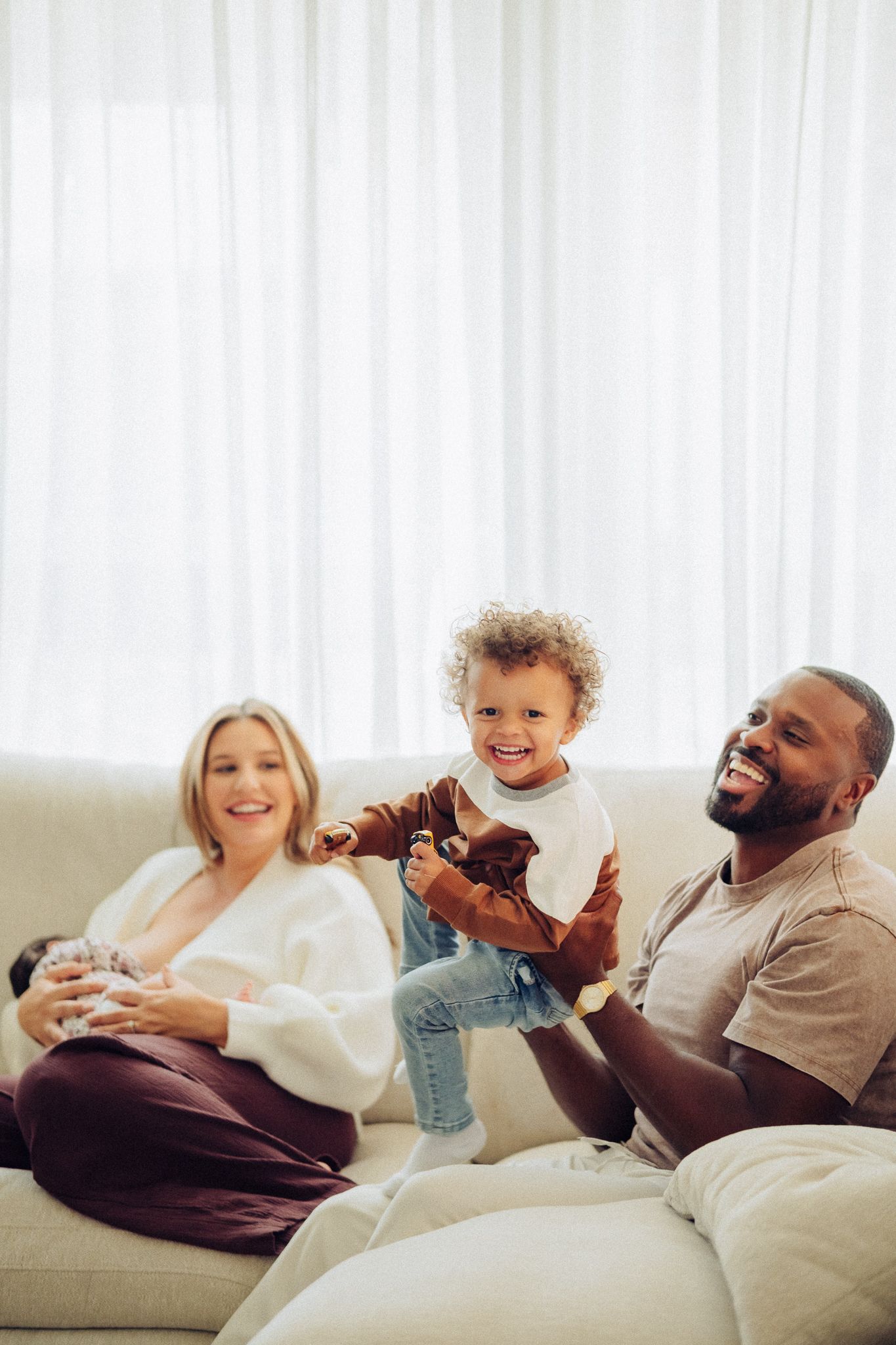 Happy interracial family relaxing on a couch, with a mother breastfeeding, father lifting a curly-haired toddler.