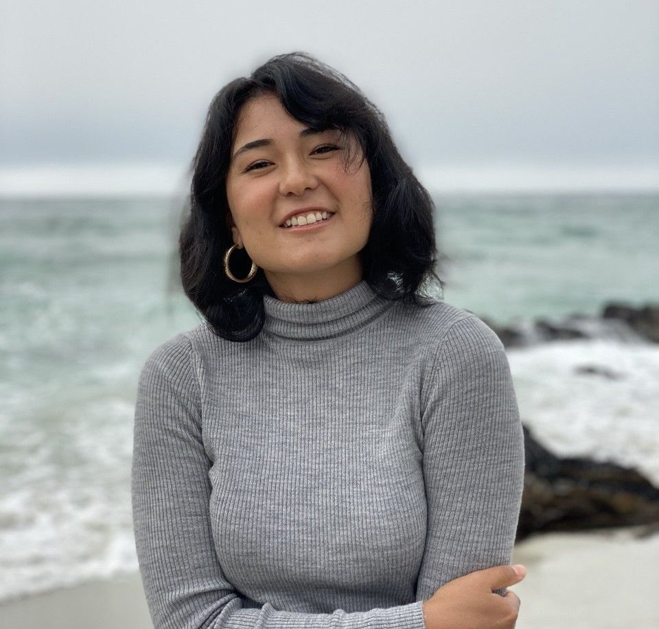 Joyful person in a gray turtleneck sweater standing by a rocky beach on an overcast day with ocean waves in background.