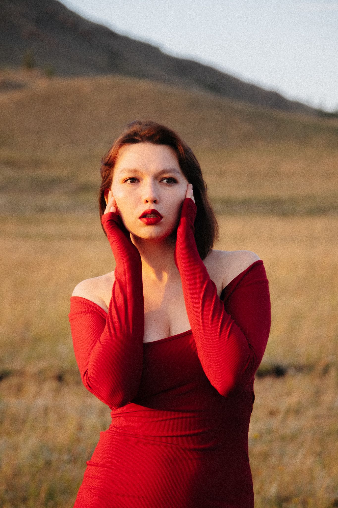 A dramatic fashion portrait in a red dress and gloves against a golden field at sunset.