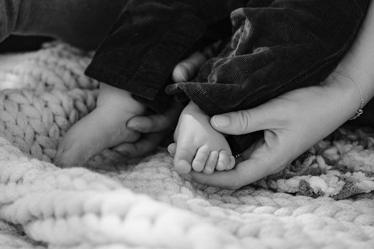 Black and white close-up of gentle hands touching tiny baby feet on a textured knit blanket.