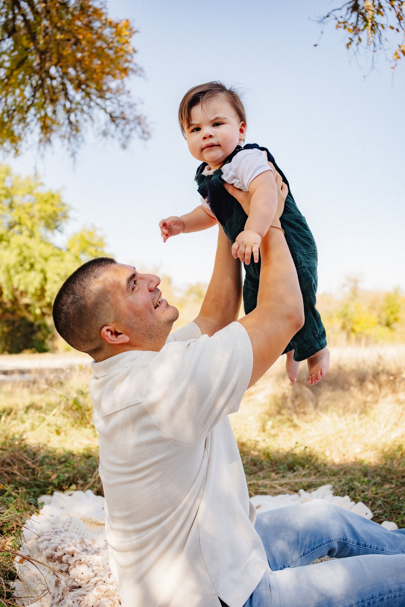 A playful moment captured outdoors as a parent lifts a small child wearing overalls against an autumn backdrop.