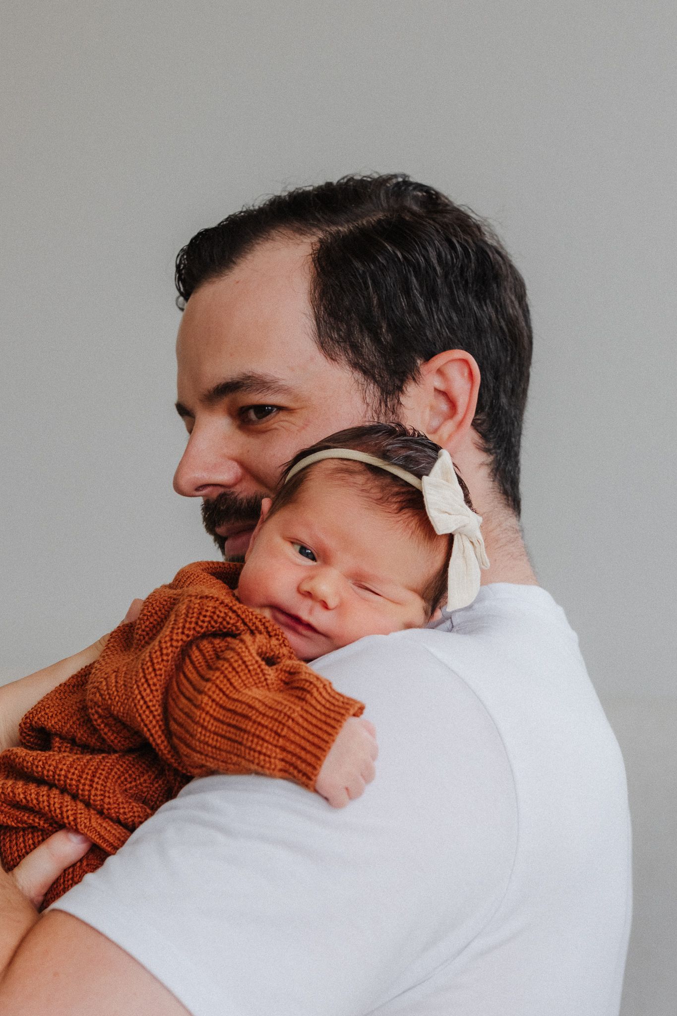 A bearded man in a white shirt tenderly holds a sleeping newborn baby girl wearing an orange knit outfit and bow headband.