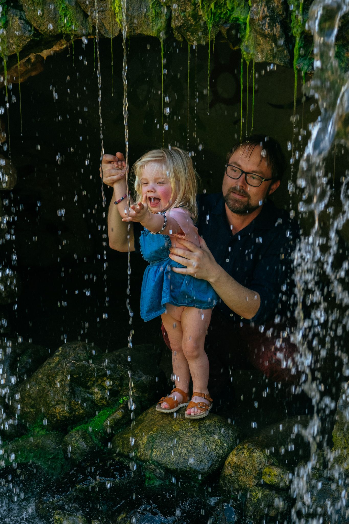 A man holds a laughing toddler girl in a blue dress behind a waterfall, standing on mossy rocks.