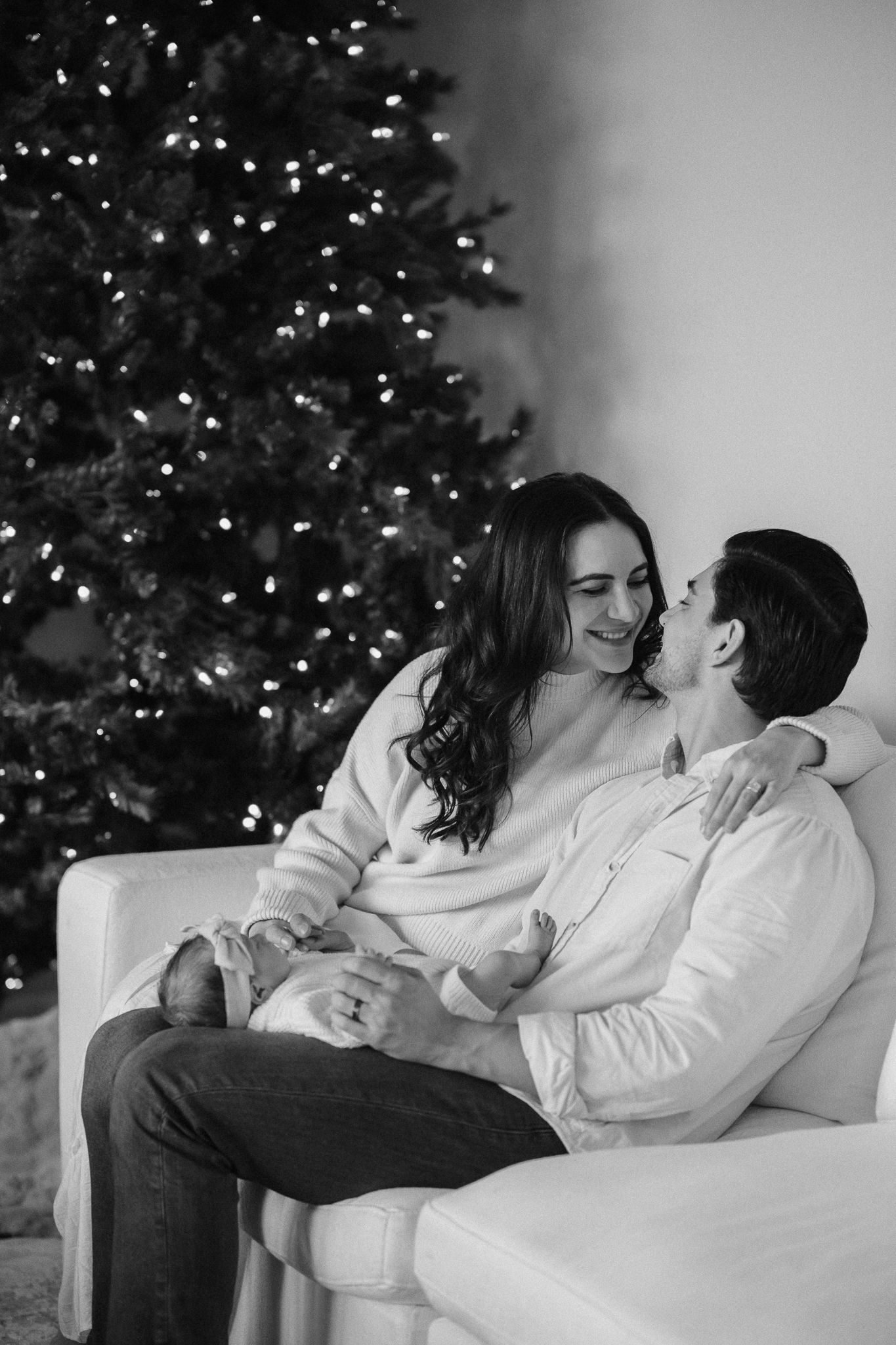 Black and white photo of a couple sitting on a sofa, smiling and embracing in front of a lit Christmas tree.