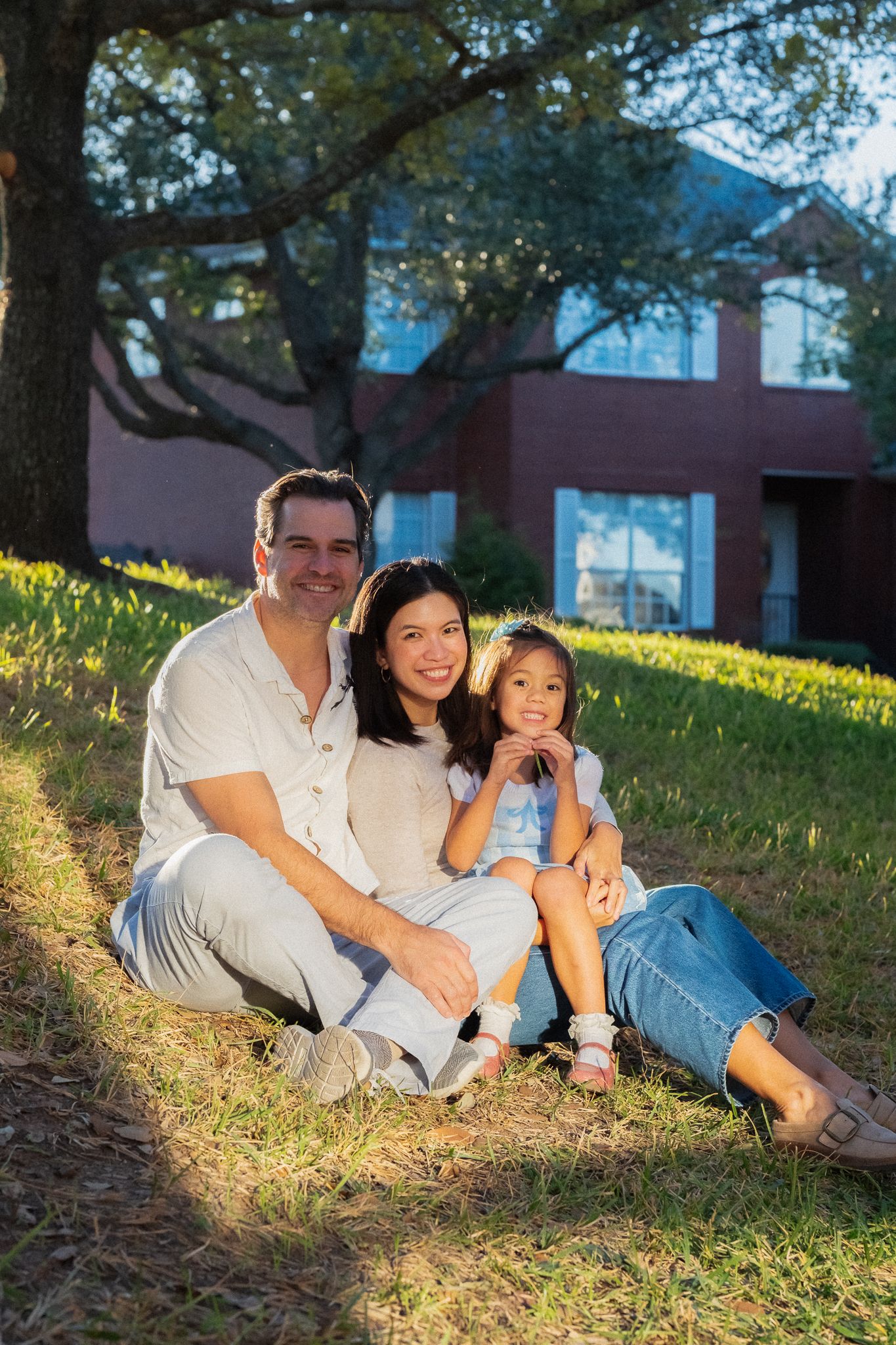 Family relaxing together on a sunny hillside under trees near a brick building.