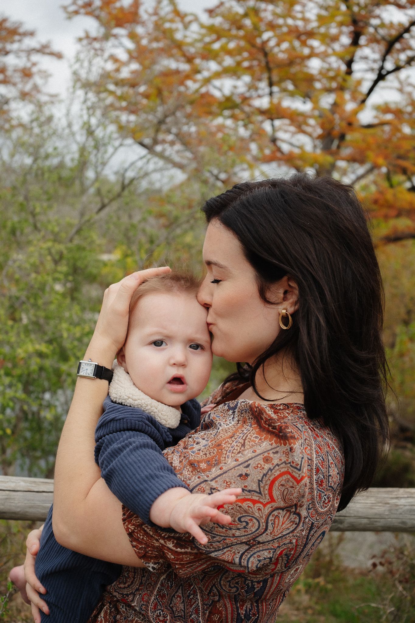 A tender moment captured outdoors with a baby in navy blue being kissed against a backdrop of autumn foliage.