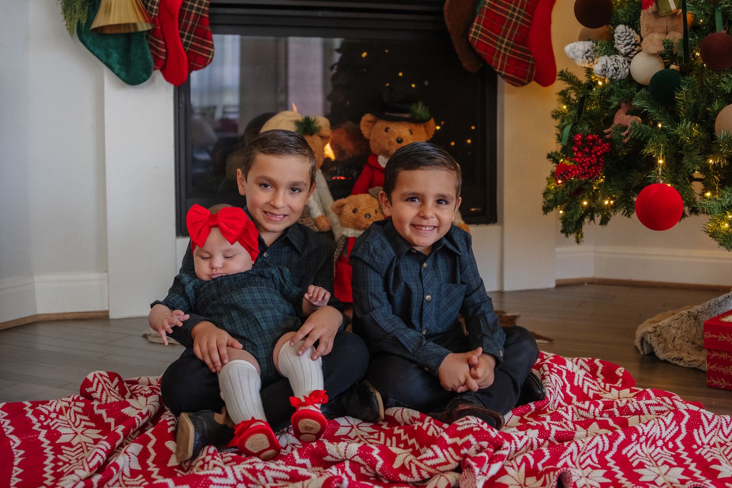 Three children sit on a red and white patterned blanket in front of a decorated Christmas tree with stockings hanging above.