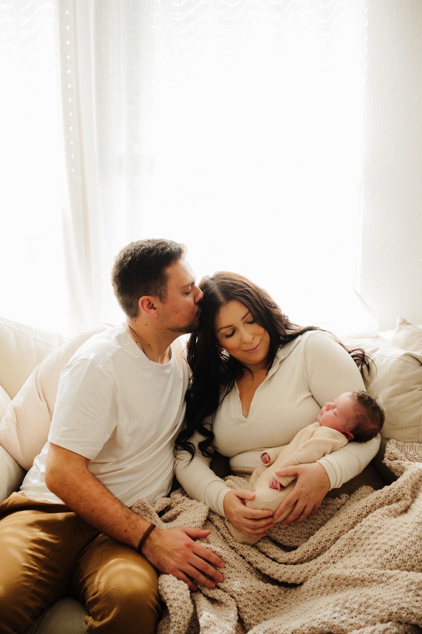 Family lying together on bed with knitted blanket in warm sunlit room showing progression of tender moments.