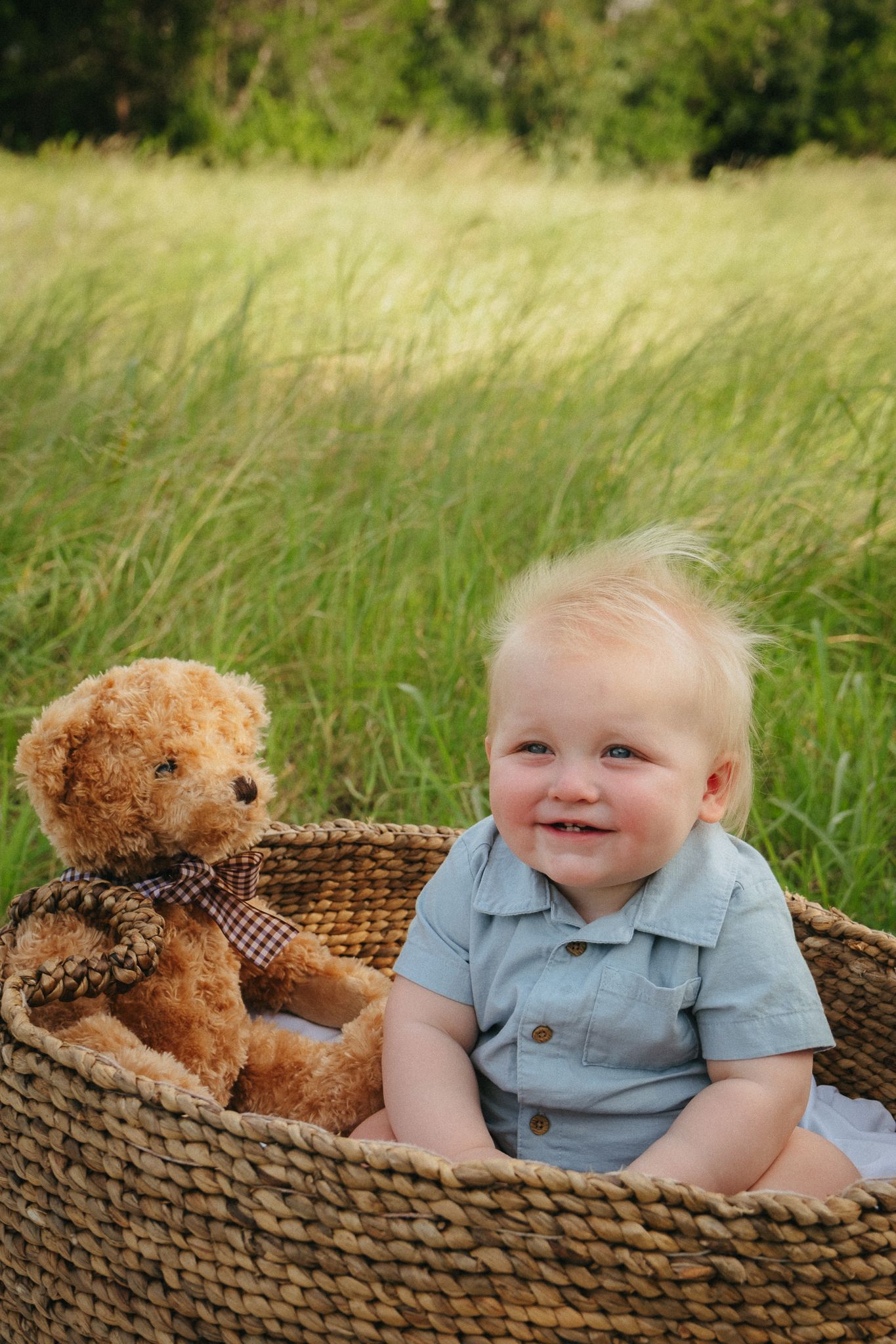 A smiling baby sits in a woven basket with a brown teddy bear against a backdrop of tall grass.